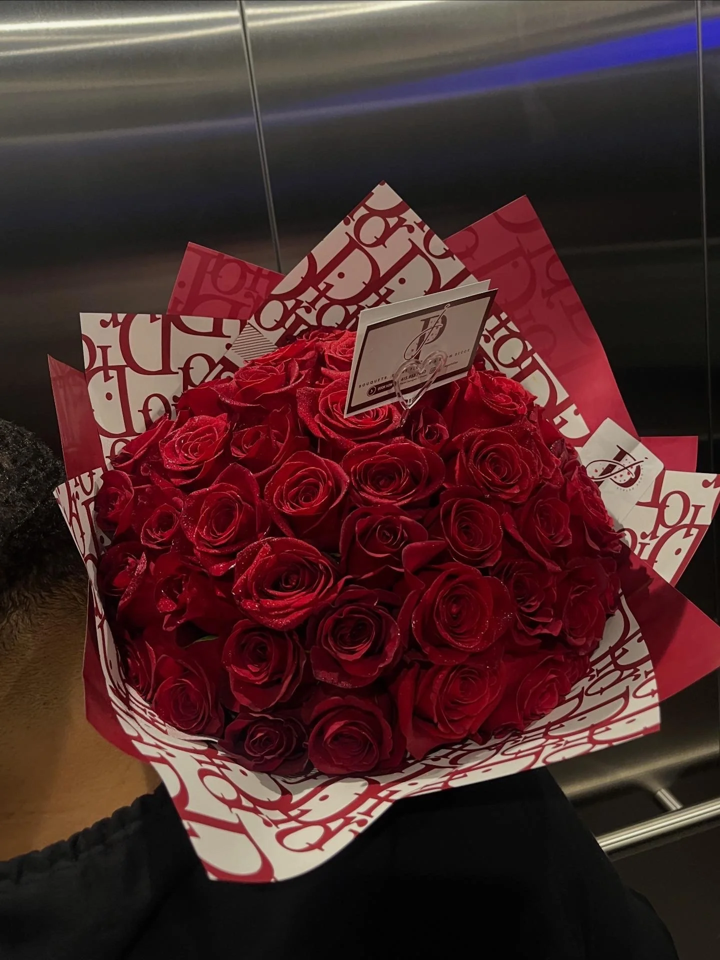A bouquet of red roses wrapped in decorative paper with white and red patterns. A card is inserted among the roses, displaying elegant script and a logo.