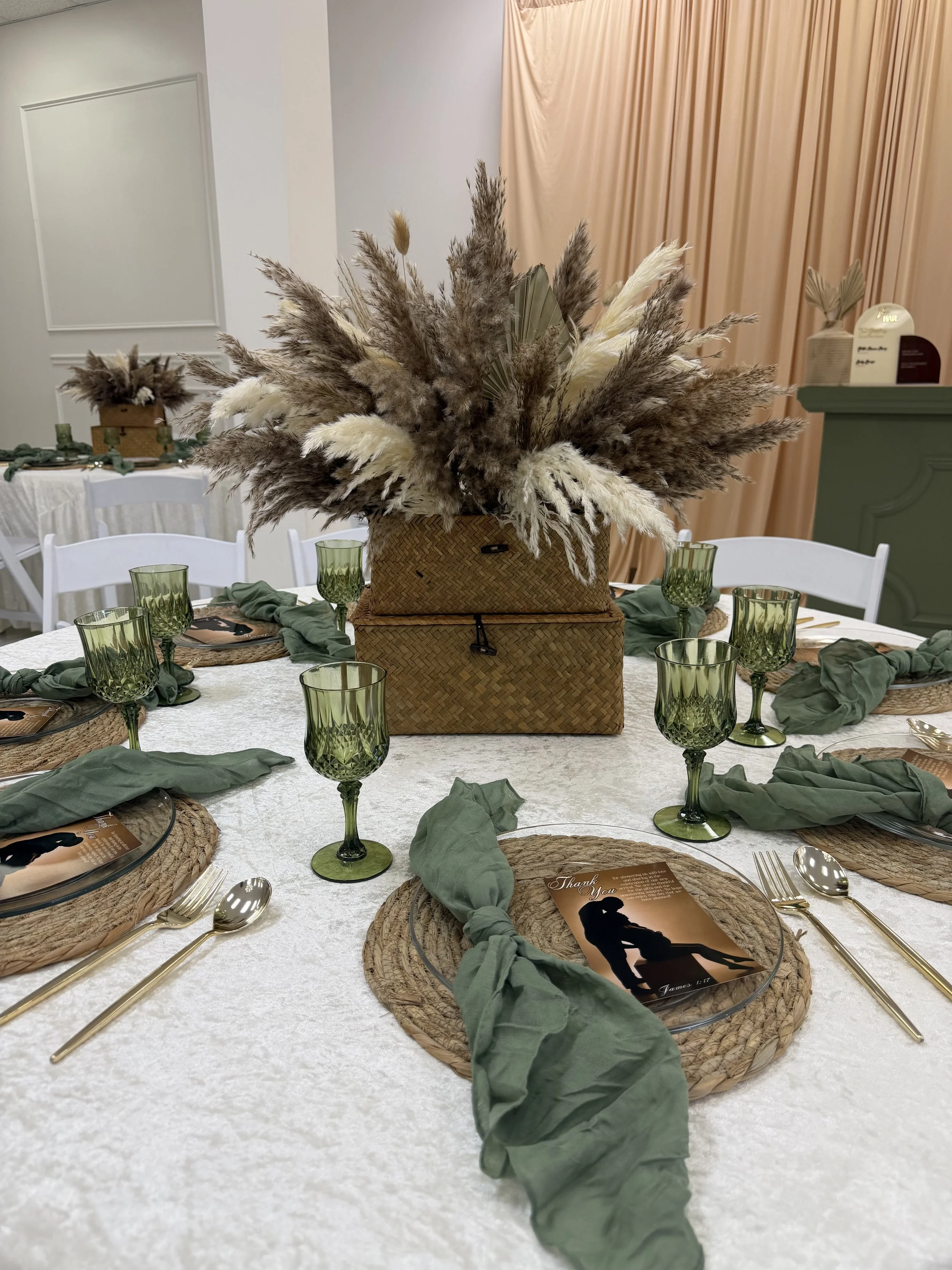 A round table decorated with a large floral arrangement of pampas grass, surrounded by white chairs, green glasses, and place settings with woven placemats and green napkins. A thank you card with an image of a person and Bible verse is on each place