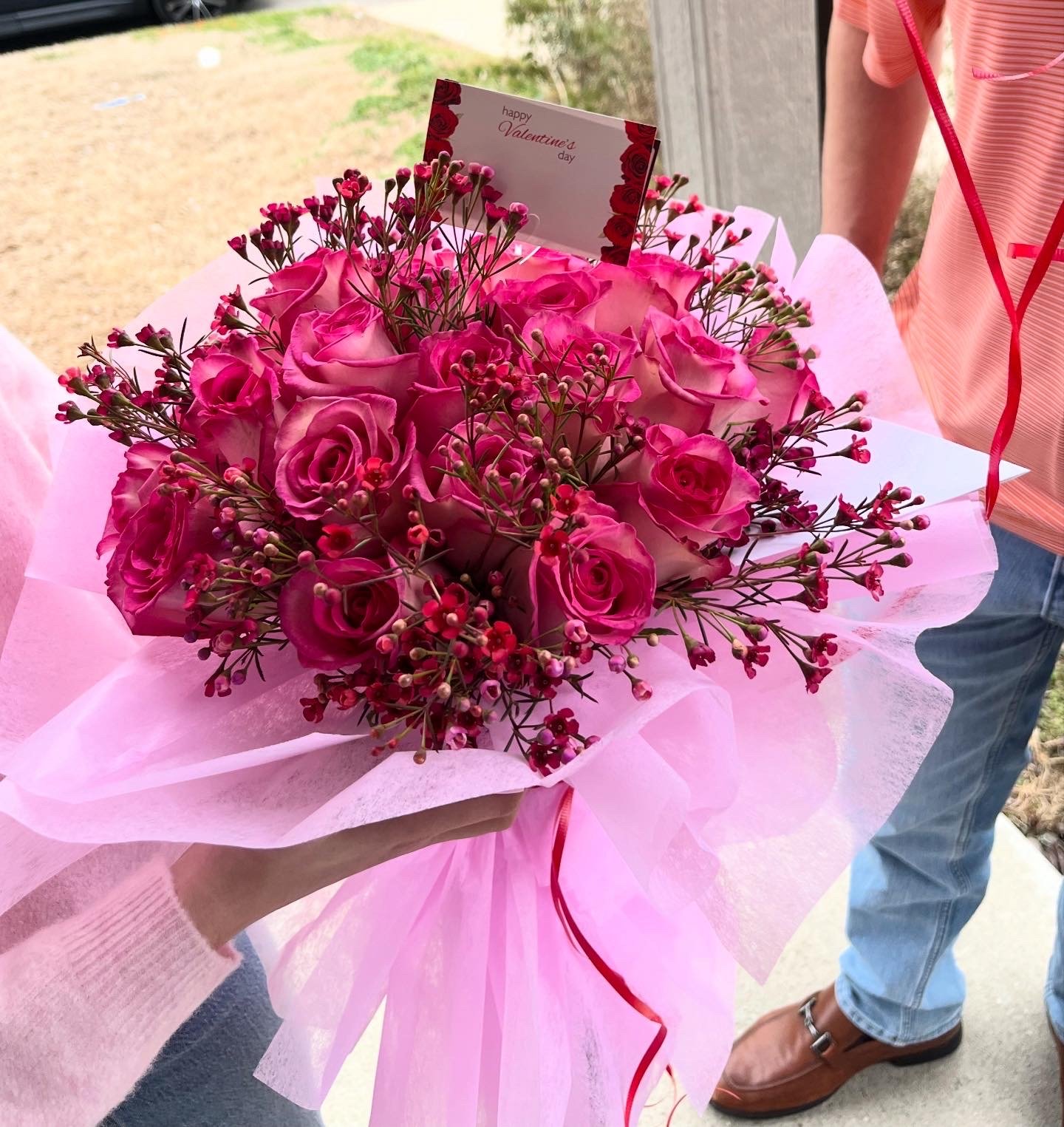 A person holding a pink bouquet of roses and small pink flowers with a 'Happy Valentine's Day' card.