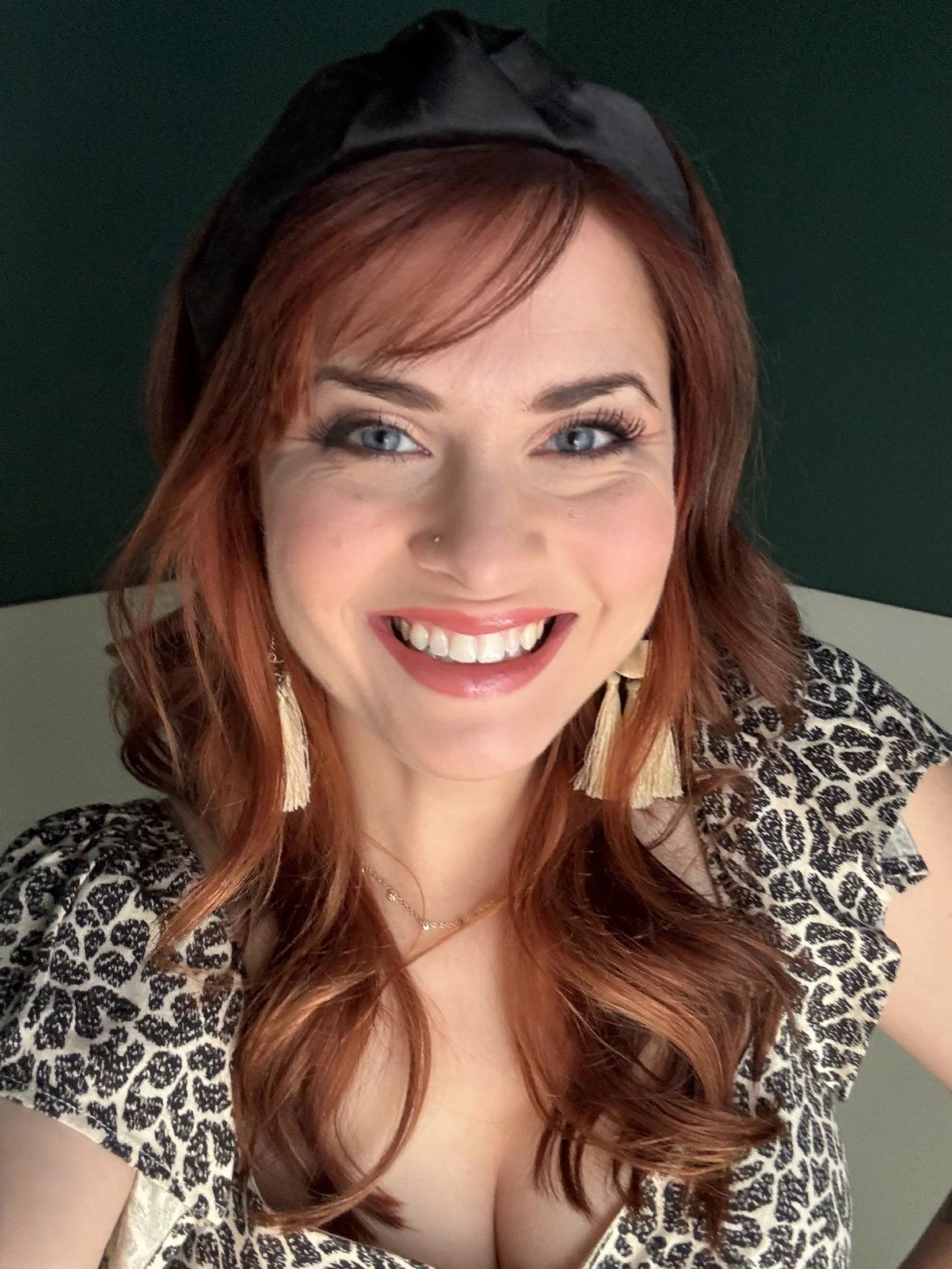 A woman with red hair, blue eyes, wearing a black headband, earrings, a necklace, and a leopard print top, smiling.