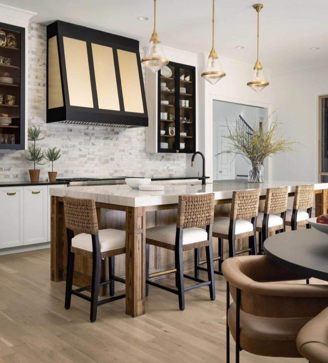 Modern kitchen with white island, four woven barstools, black and beige cabinetry, three pendant lights, a vase with tall branches, and decorative items on shelves.