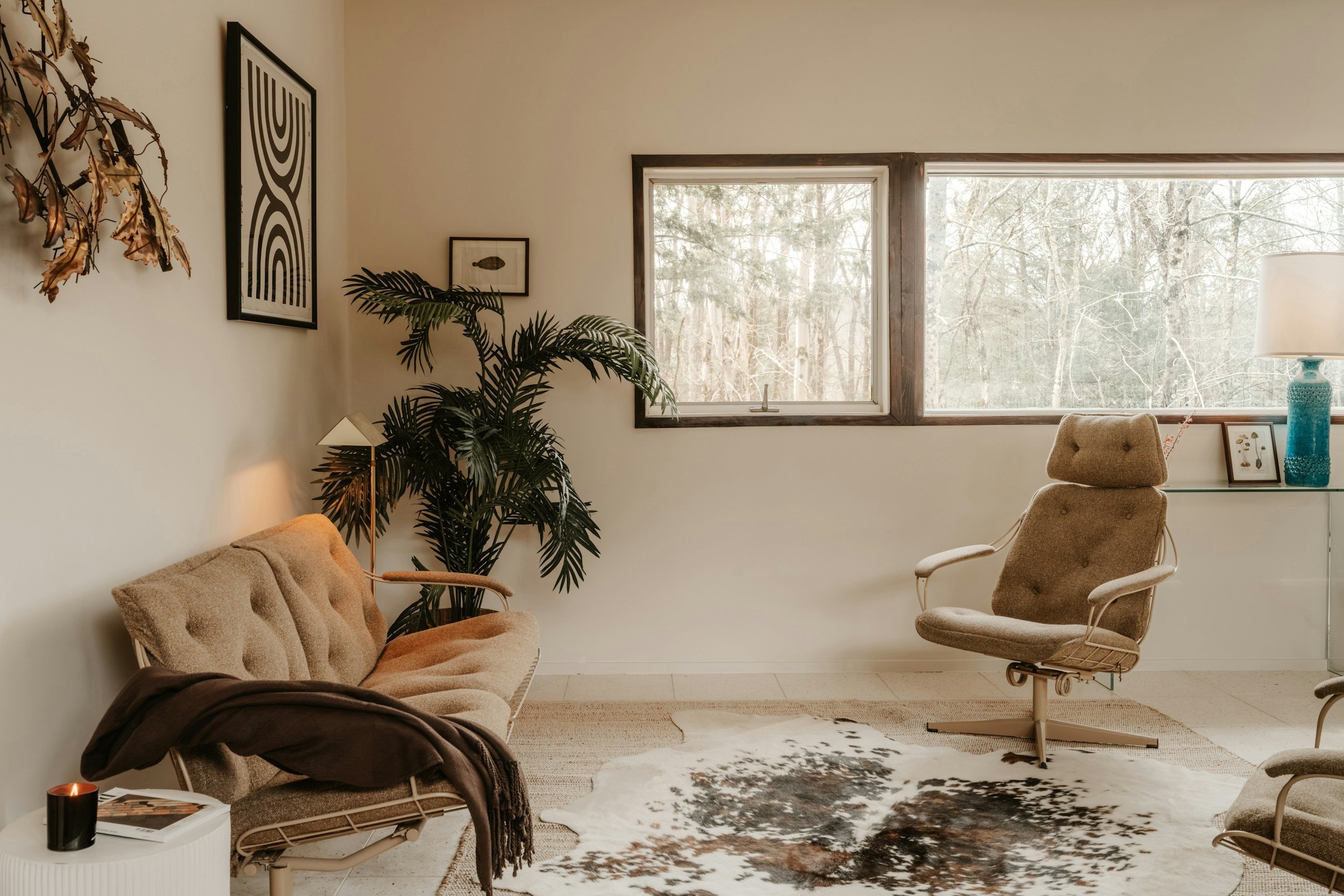 A cozy living room with beige upholstered chairs, a cowhide rug, and a large window overlooking a wintery wooded scene. Decor includes framed art, a tall turquoise lamp, and indoor plants.