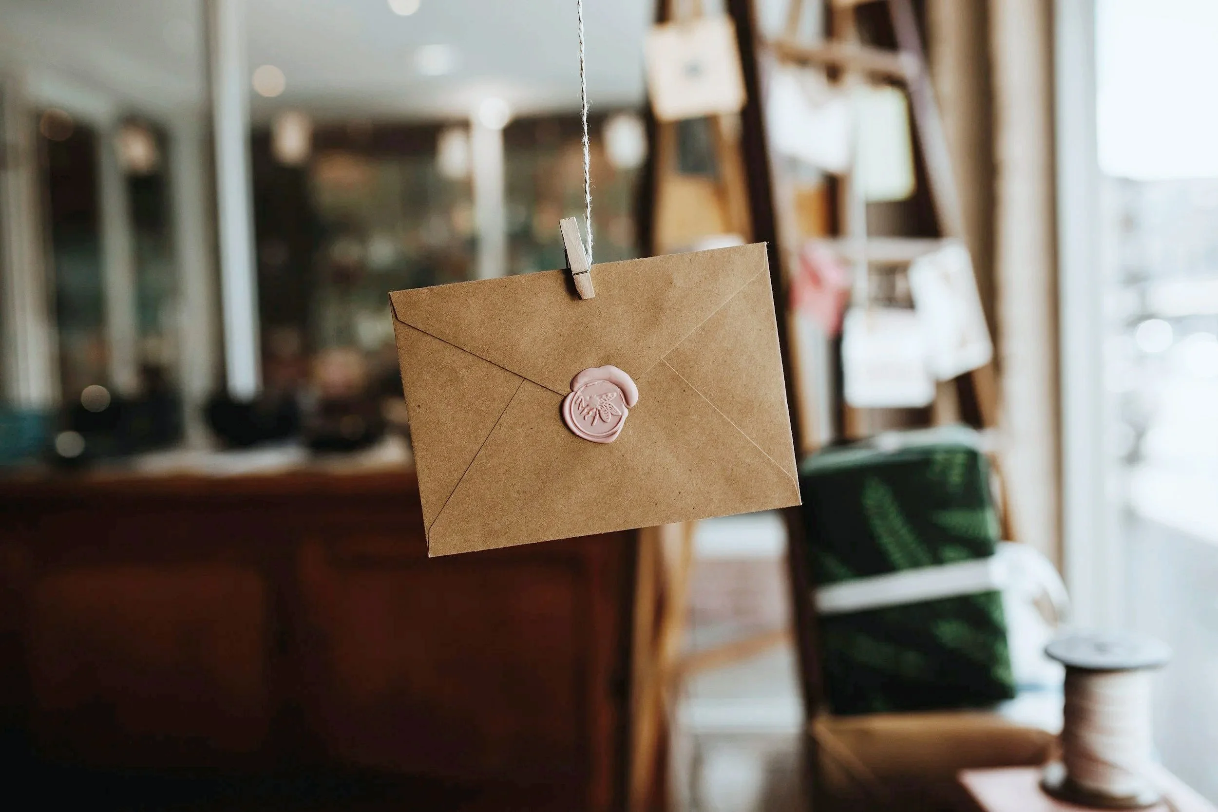 A brown envelope with a pink wax seal hanging from a string inside a cozy shop or cafe, with shelves and a window in the background.