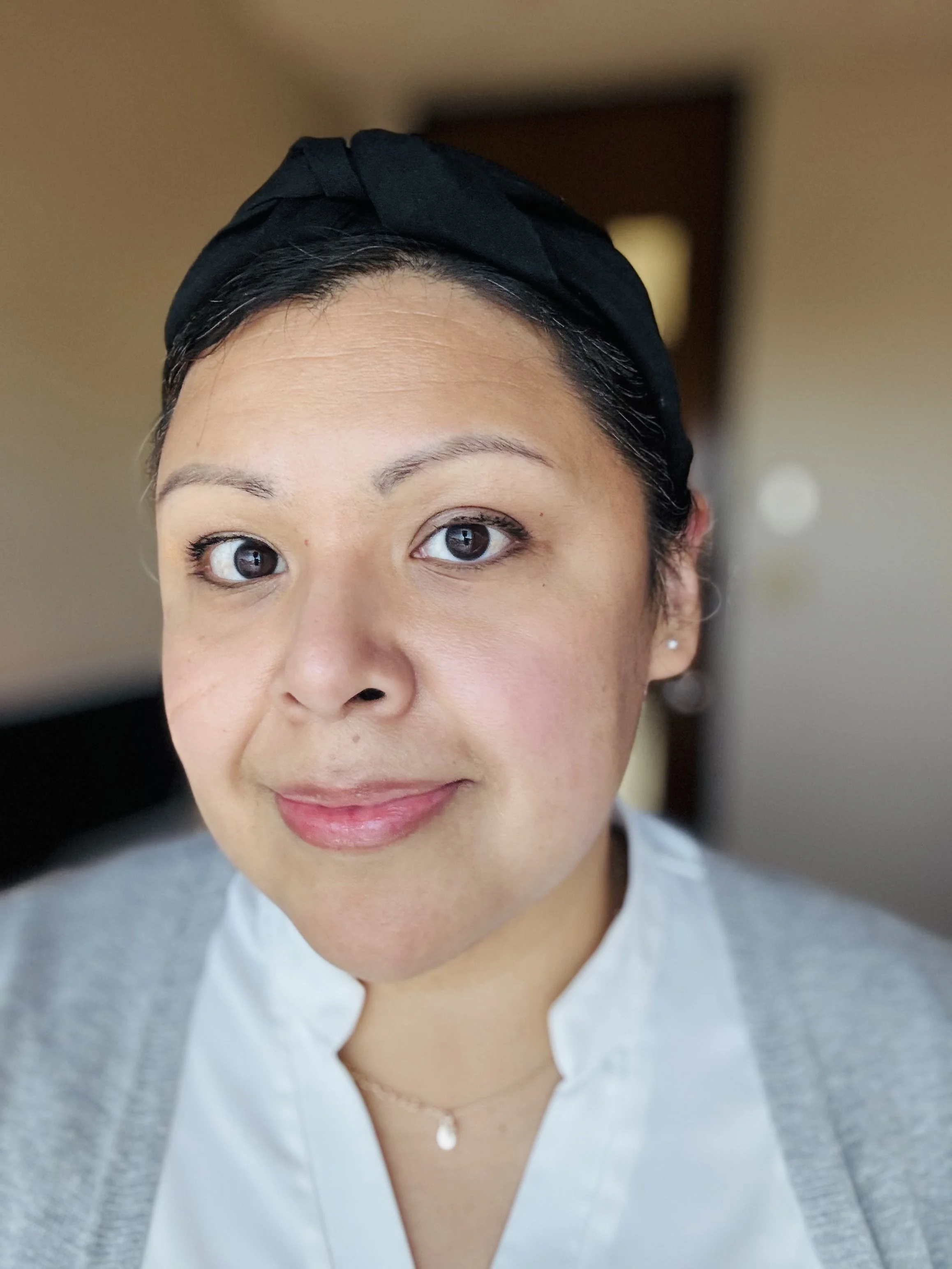 Close-up portrait of a woman with short dark hair in a black headscarf, wearing light makeup, earrings, a white blouse, and a delicate necklace.