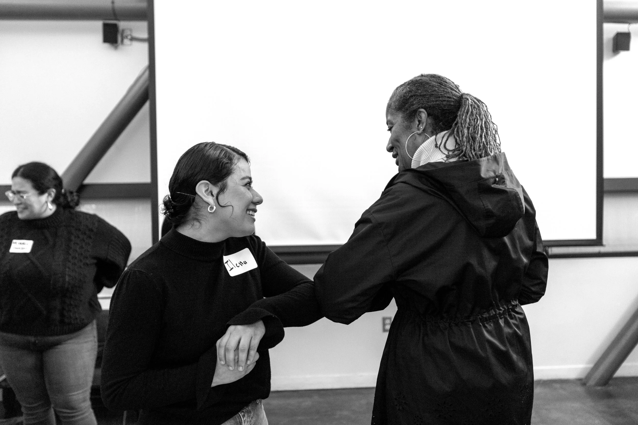 Two women smile at each other during a conversation in a room, one appears to be comforting or encouraging the other.