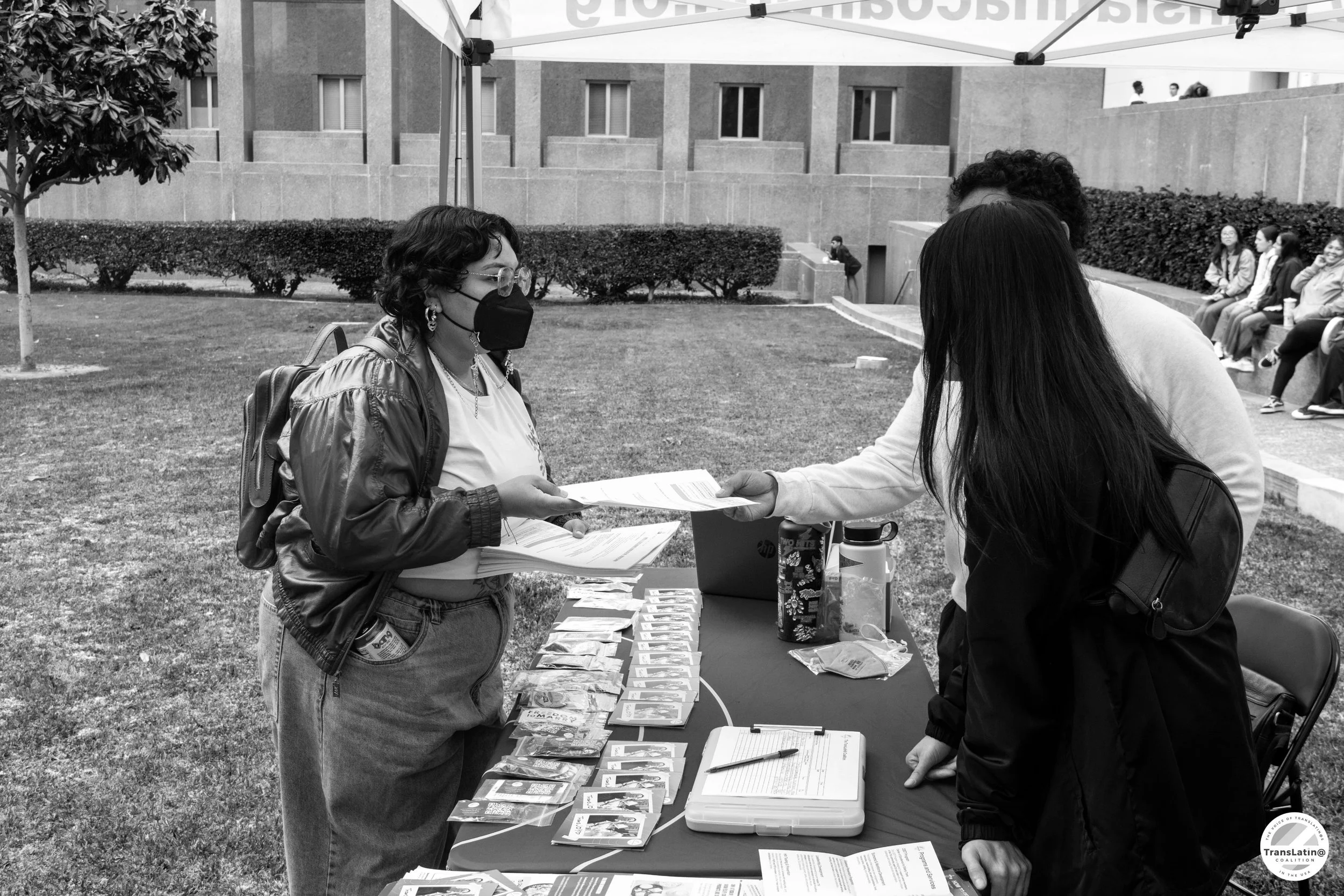 People at an outdoor event; two women exchanging papers at a table with pamphlets and flyers, some individuals seated in the background.