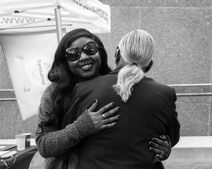 A woman with long dark hair, sunglasses, and dark nail polish hugging a person with long, light-colored hair tied back, in an outdoor setting with a canopy and a banner in the background.