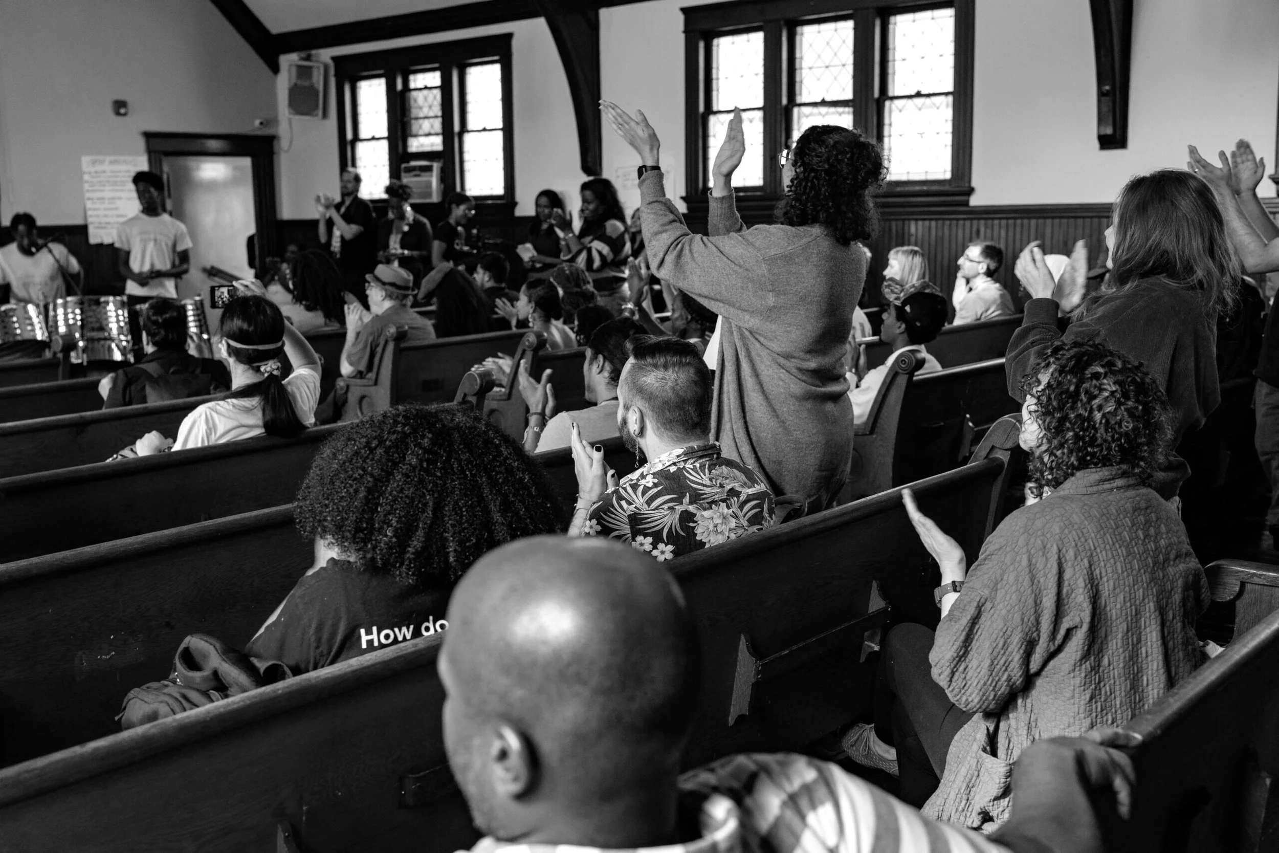 A diverse group of people sitting and standing in a church or auditorium, engaging in a communal activity, with some raising their hands and others taking pictures or clapping.