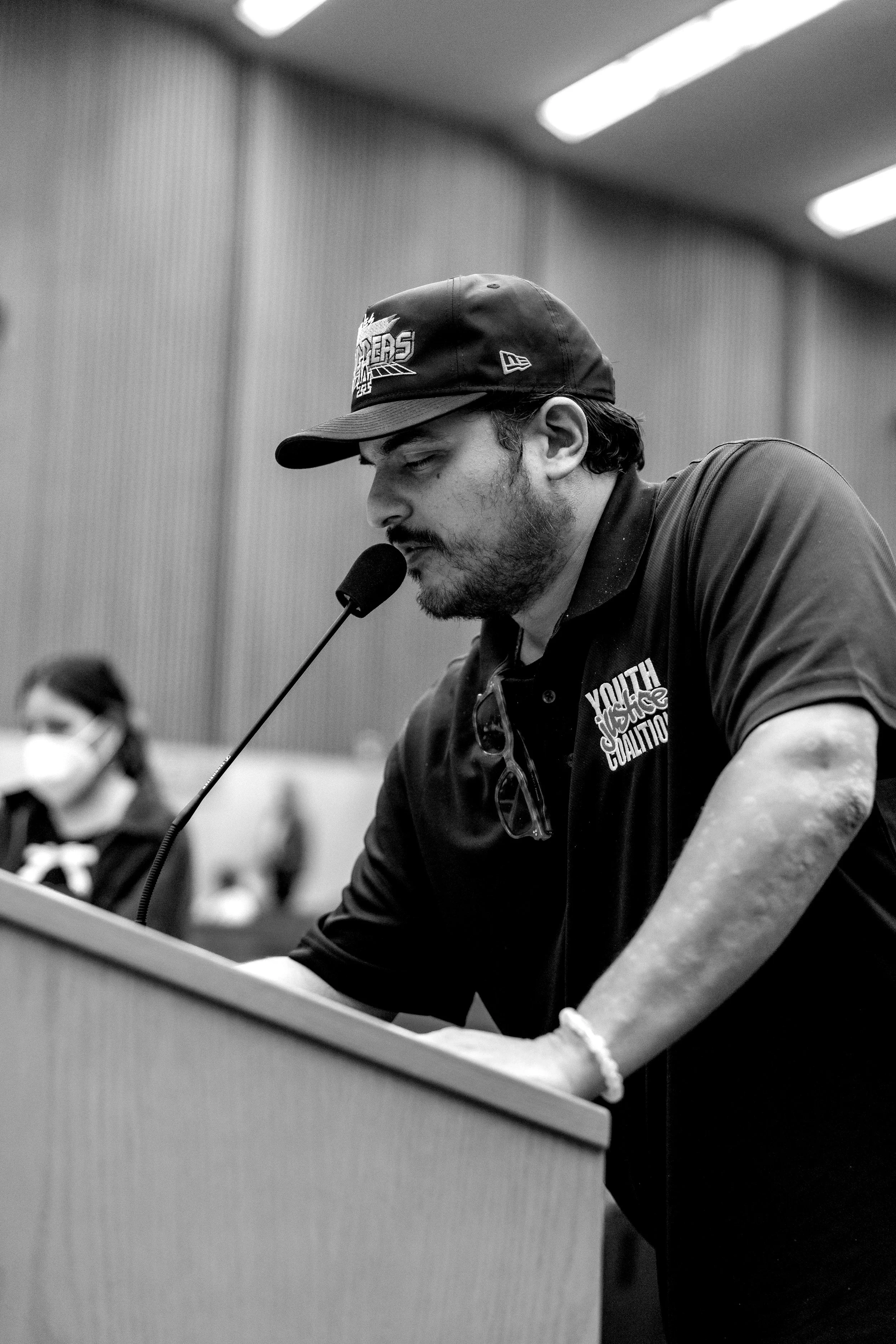 A man wearing a Los Angeles Kings cap and a Black Lives Matter shirt, speaking into a microphone at a public meeting or hearing.