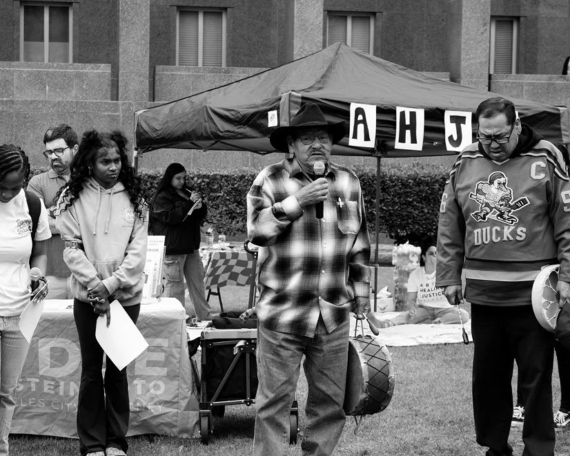 A group of people standing outdoors near a booth with a banner that has partially visible letters 'A H J.' One man in a plaid shirt and cowboy hat is speaking into a microphone, while others listen.