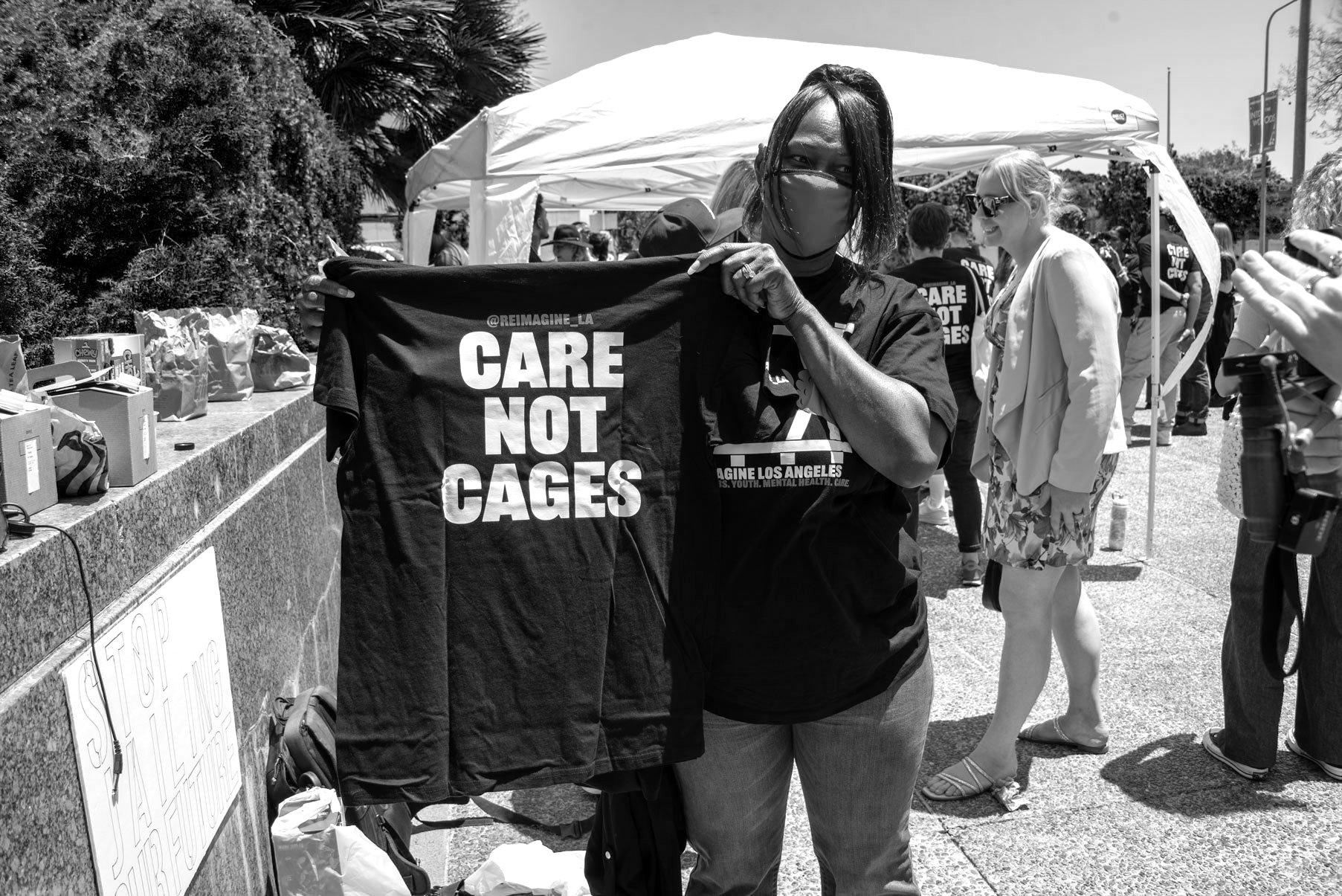 A woman wearing a face mask and dark clothing is holding a black T-shirt with the message 'CARE NOT CAGES' at an outdoor event or protest. Behind her, there are other people, some wearing shirts with the same message, and a tent.