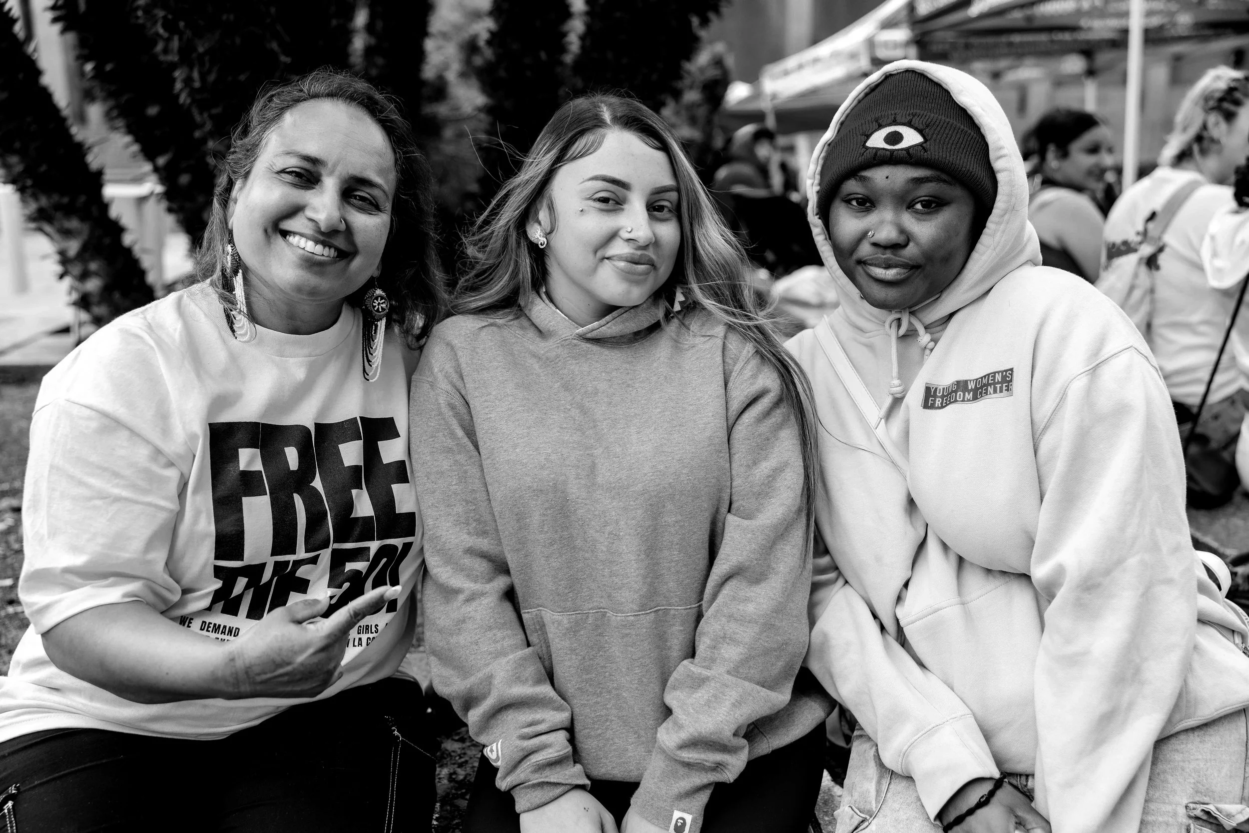 Three women sitting outdoors at a gathering or protest, smiling. The woman on the left has earrings and a nose piercing, the middle woman has earrings, a nose piercing, and curly hair, while the woman on the right wears a beanie with an eye symbol, a hoodie with a patch, and has a nose stud. There are other people and tents in the background.
