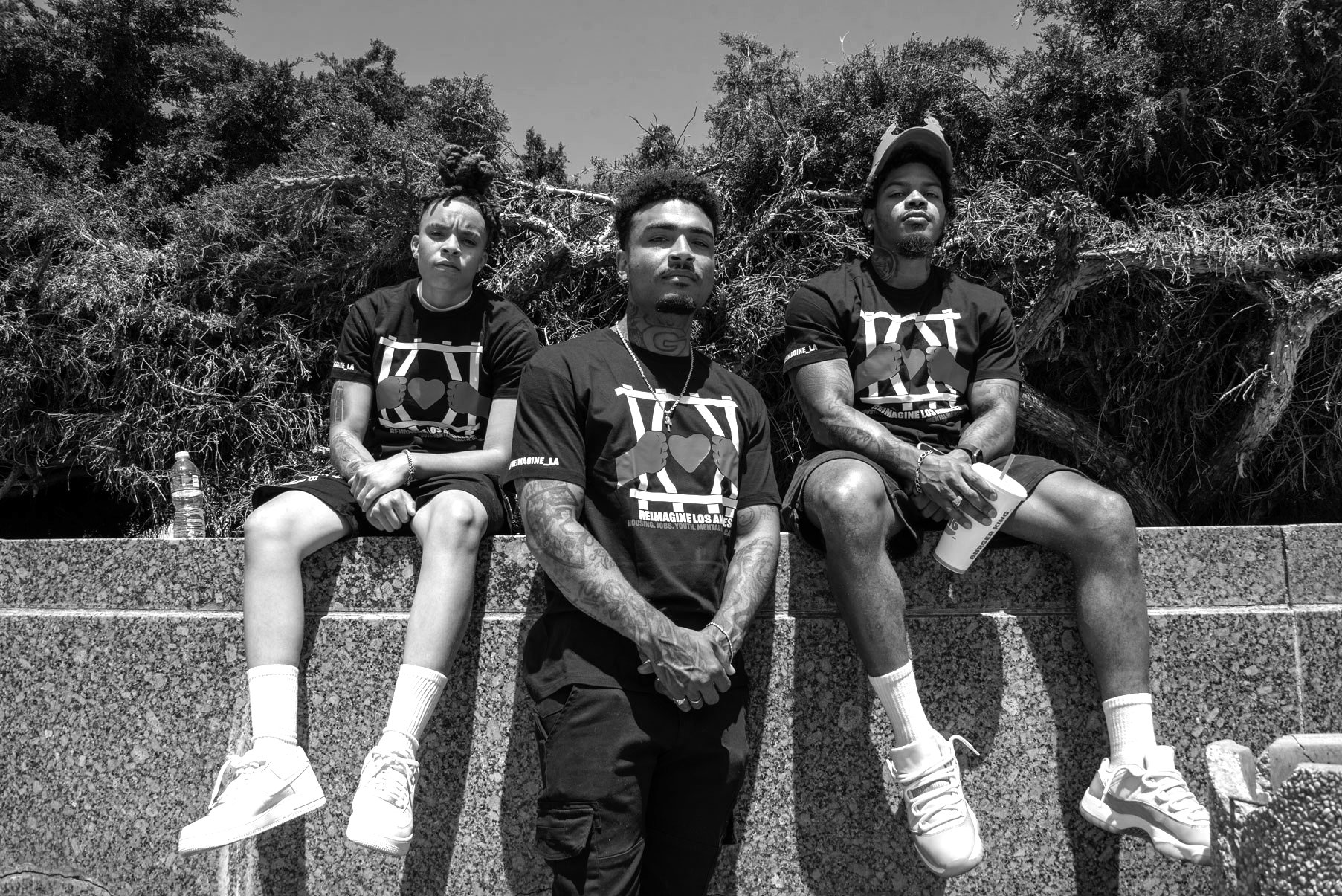 Three young men sitting on a stone ledge outdoors, wearing matching black T-shirts with a graphic of two fists holding a heart, and shorts. The man in the middle is standing and looking at the camera, the woman on the left has their hair styled in dreadlocks, and the man on the right is wearing a cap backwards. There is a water bottle next to the woman and a drink cup with a straw with the man on the right. Behind them is a large bush or tree with twisted branches.