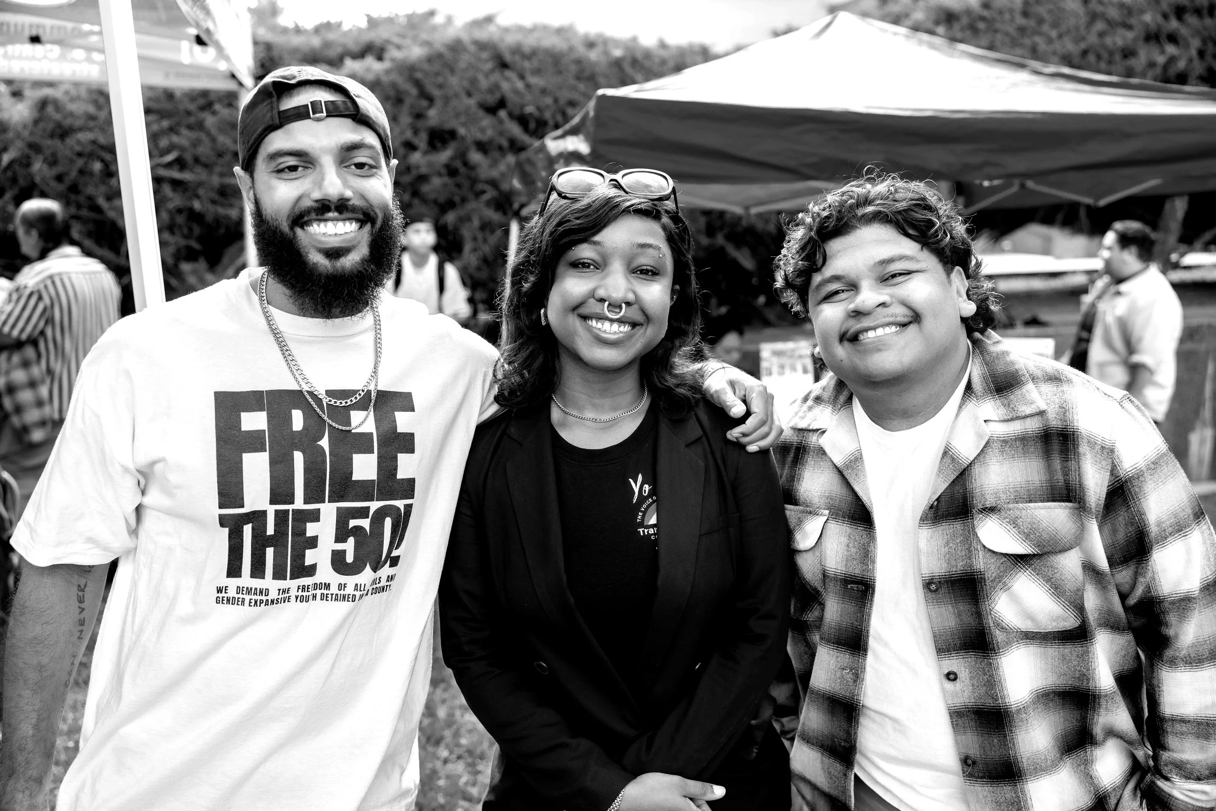 Three people smiling and standing close together at an outdoor event, with tents and other people in the background.