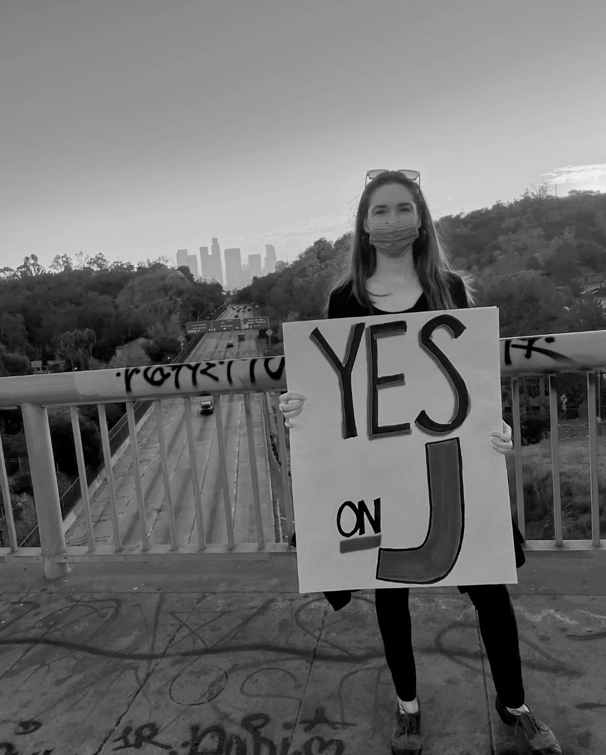 A woman wearing a face mask holding a sign that says 'YES on J' stands on a bridge over a highway with a city skyline in the background.
