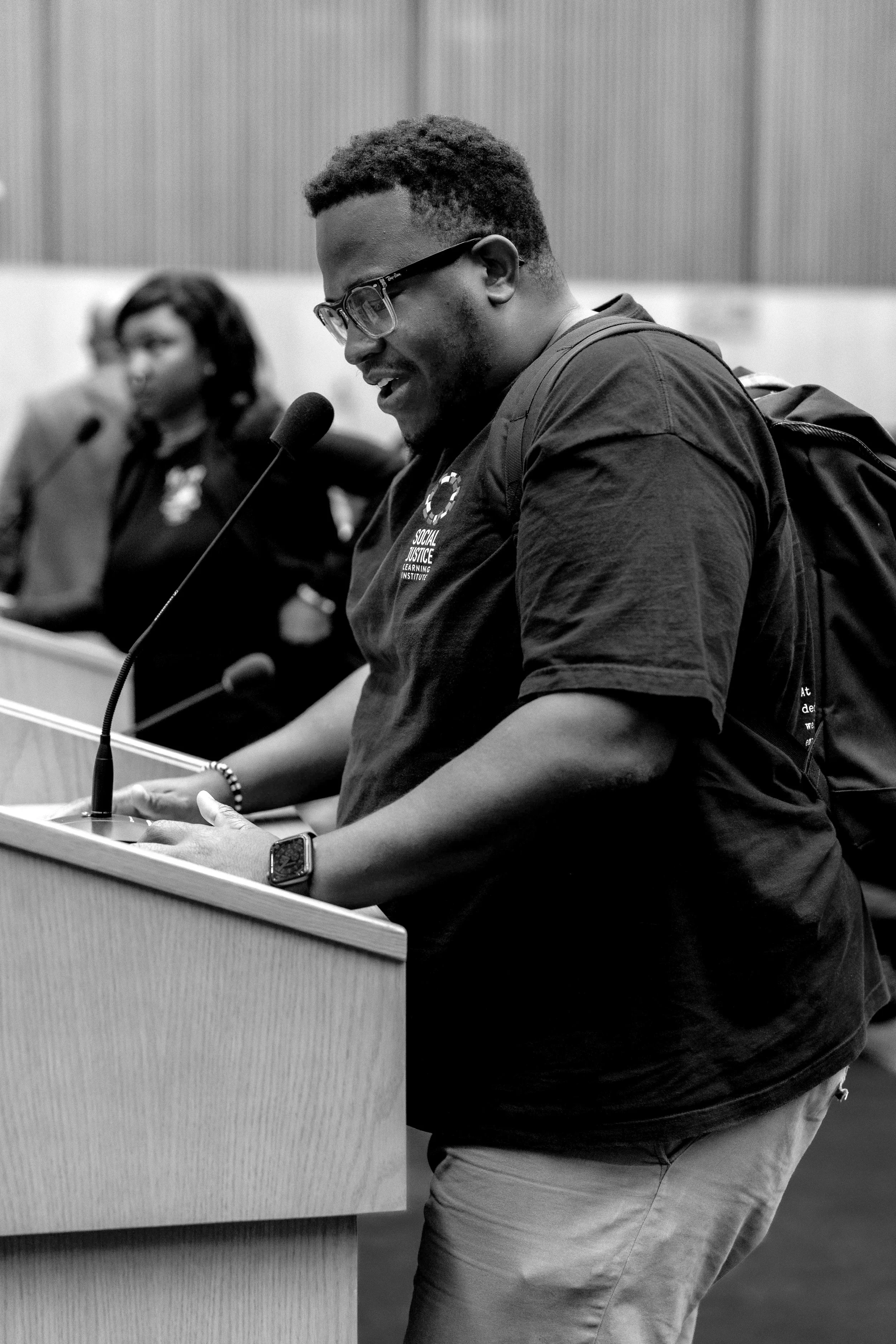 A man wearing glasses and a T-shirt reading 'Social Justice Learning Institute' speaking into a microphone at a panel or conference, with a woman sitting behind him also at the panel.