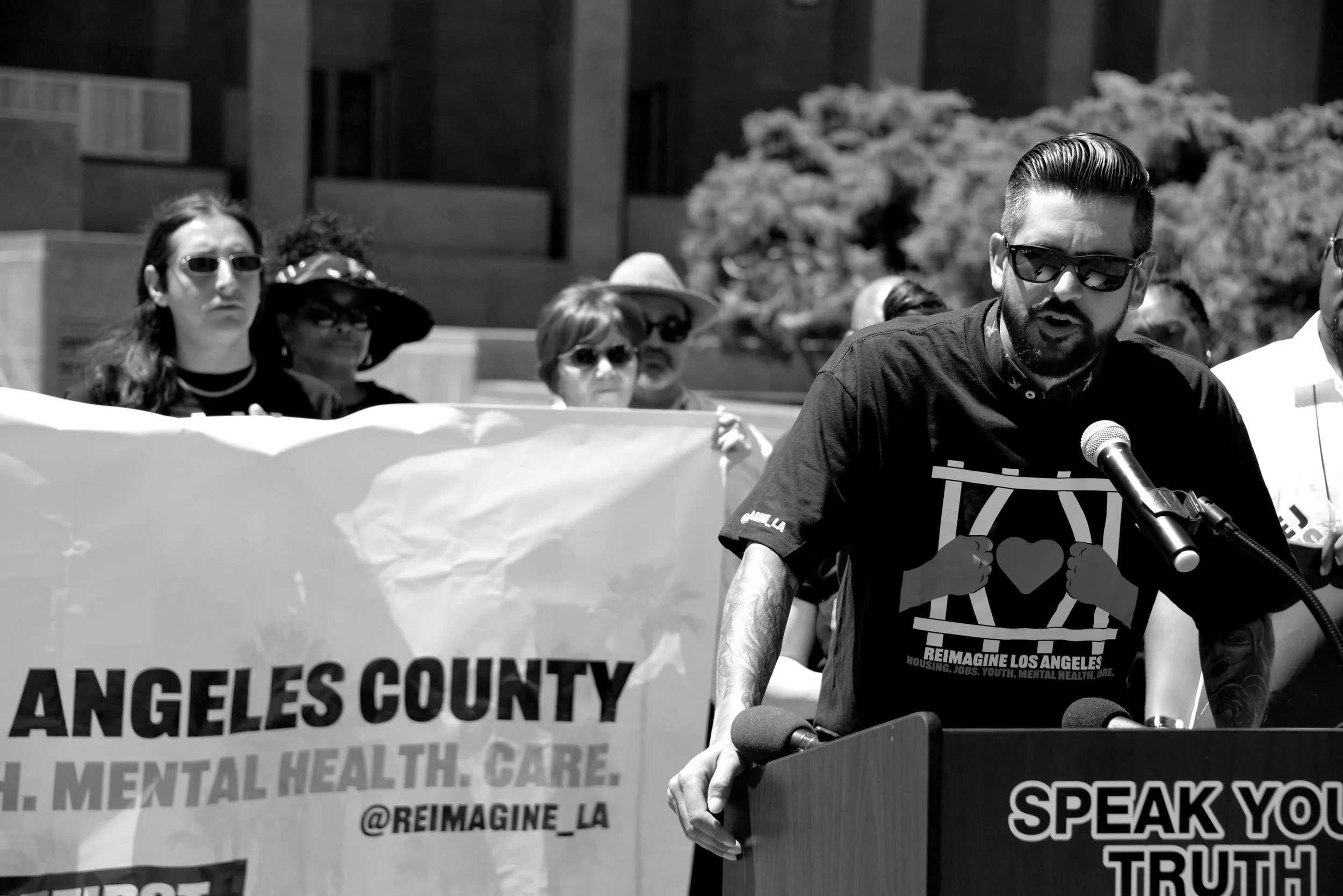 A man with dark hair, sunglasses, and a beard speaking at a podium during a protest or rally. He is wearing a black t-shirt with a peace symbol and text. Behind him, there are several women holding a banner that reads "ANGELES COUNTY" and other partially visible words. The background shows trees and a building.