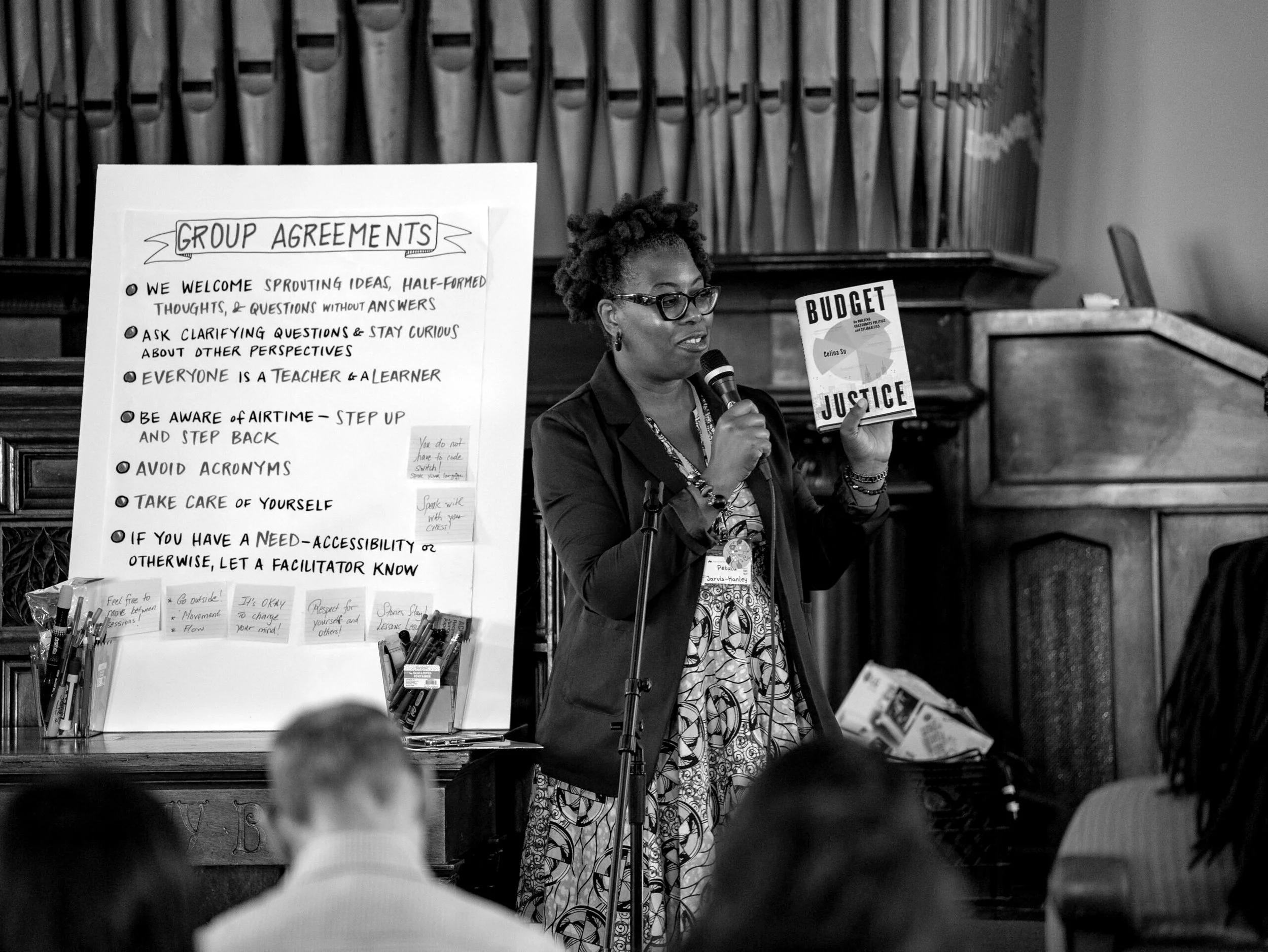 A woman with glasses holding a microphone in one hand and a booklet labeled 'Budget Justice' in the other, speaking at a presentation. There is a large poster board with handwritten notes on group agreements, and the setting appears to be a formal hall or classroom with audience members visible.