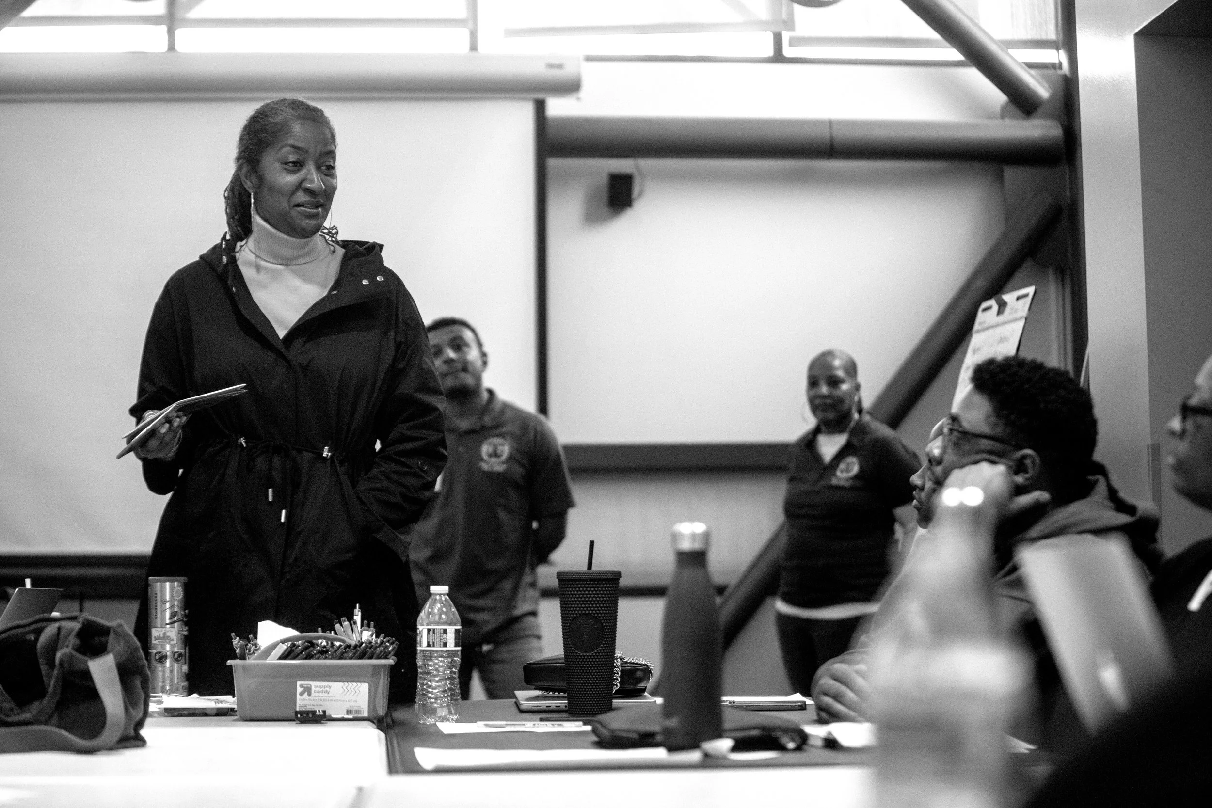 A woman is standing at the front of a group in a classroom or meeting room, speaking or presenting to seated attendees. Several bottles and educational supplies are on the table in front of her. The room has large windows at the back and a staircase on the right.