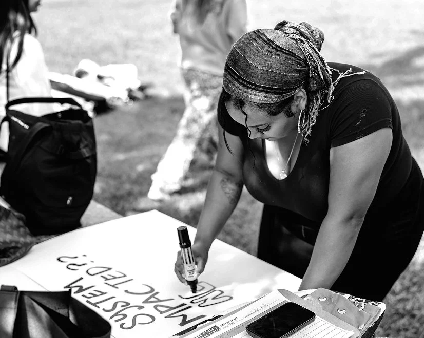 Woman with braided hair and a headscarf writing on a poster at an outdoor event.