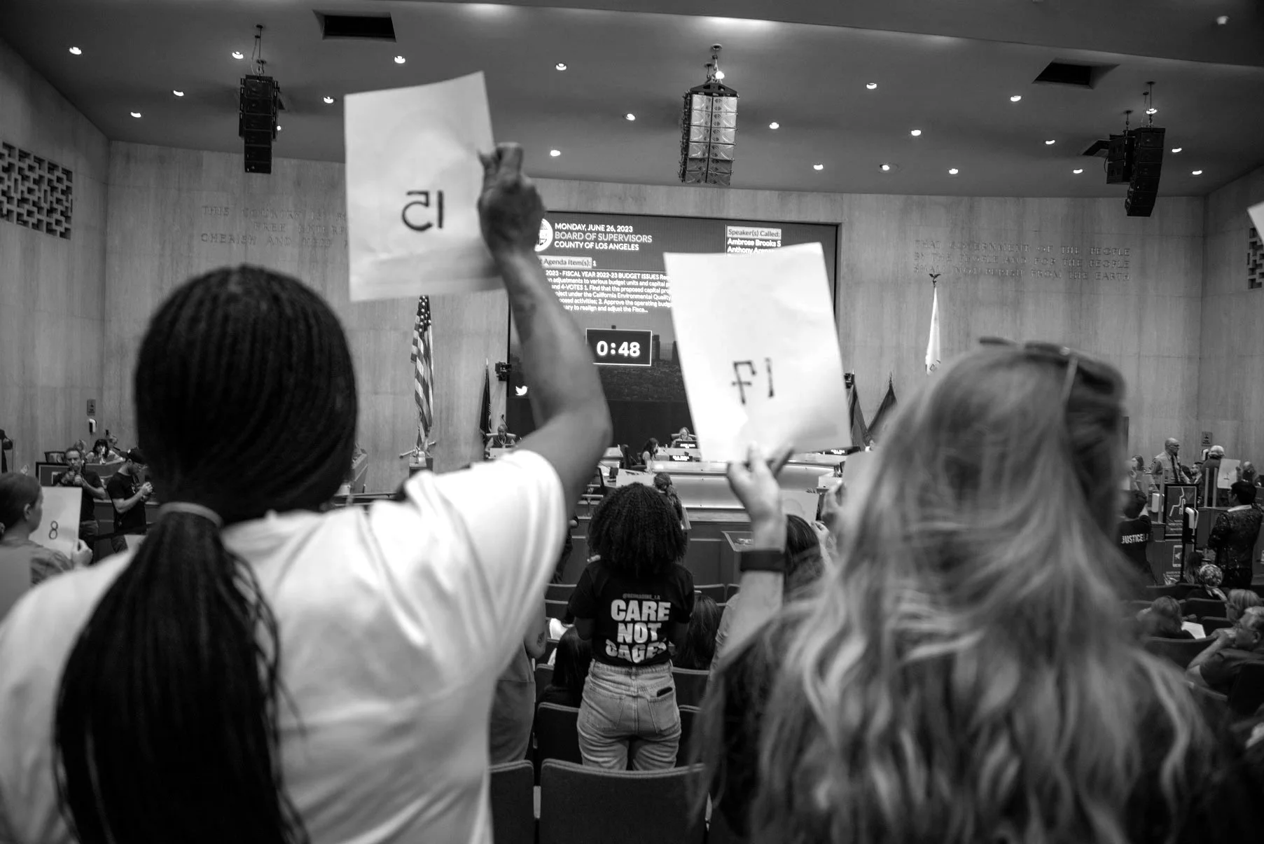 A black and white photo of a public meeting or city council session, with several people seated and some standing. A person in the foreground is holding up an election ballot, and there are large screens and flags in the background.