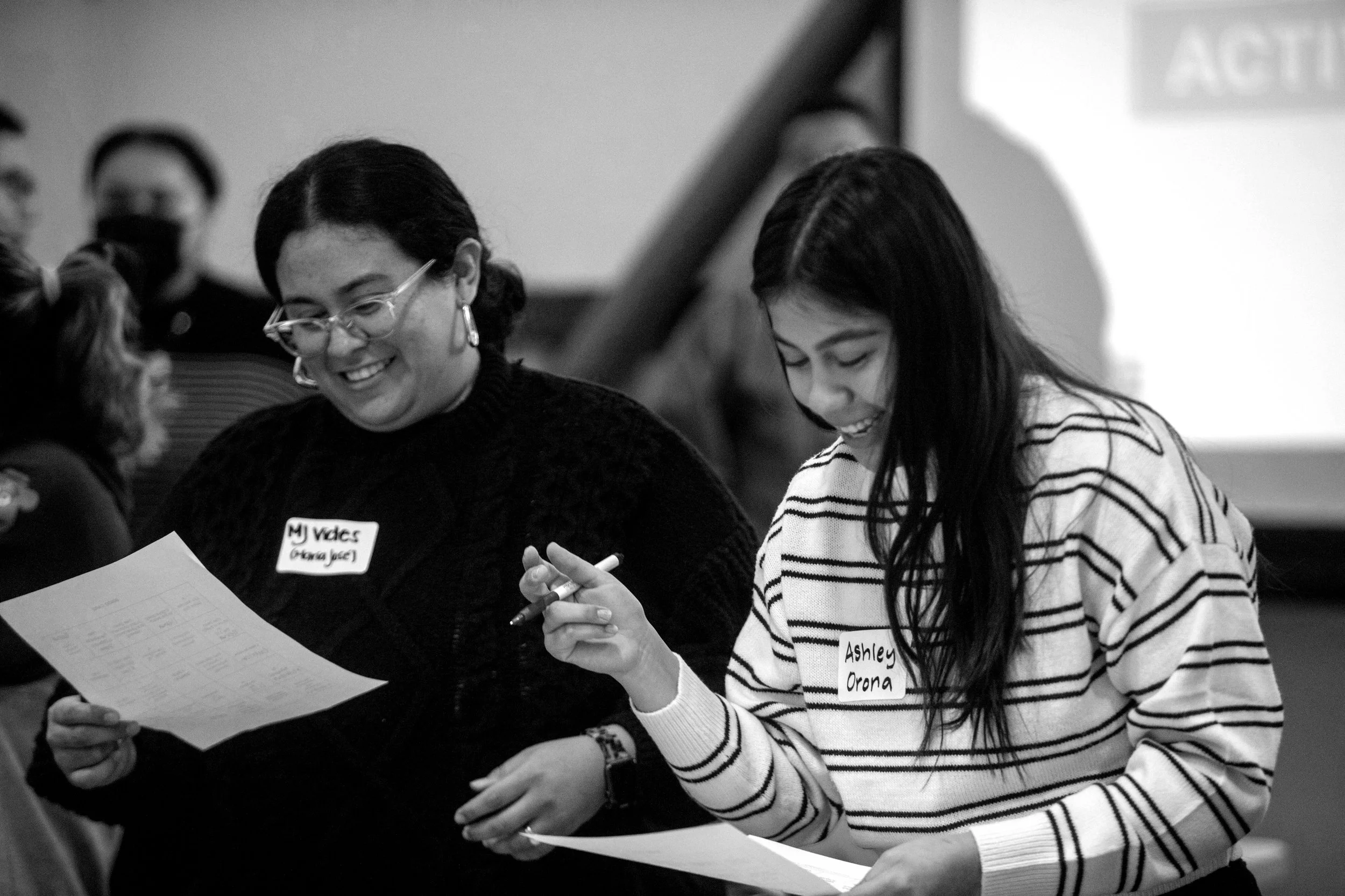 Two women, one with glasses and a name tag labeled 'MJ Vdes' and another with a striped sweater and a name tag labeled 'Ashley Orona,' are smiling and looking at papers together at a table.
