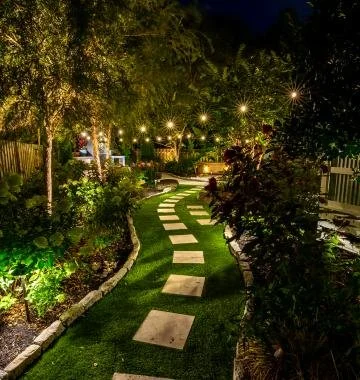Nighttime garden with a winding stone pathway illuminated by string lights, surrounded by lush greenery and plants.