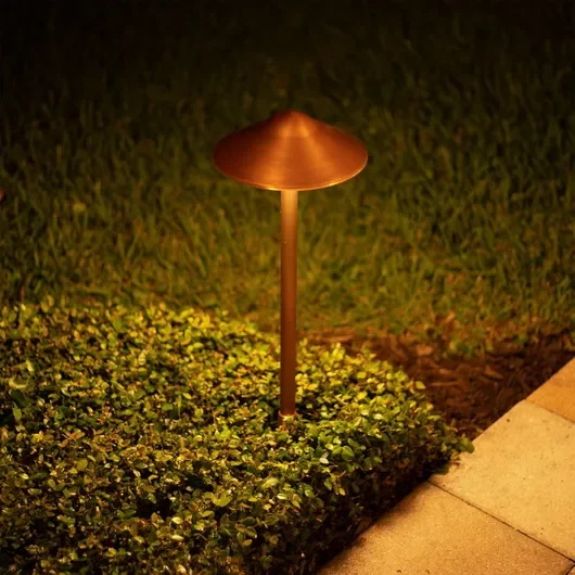 A garden light with a copper-colored shade illuminating a bed of small green plants next to a concrete pathway at night.