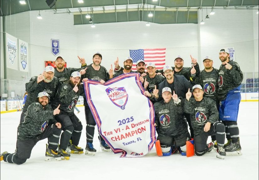 Hockey team celebrating with a championship banner on the ice rink, holding a banner that reads "2025 Tier VI-A Division Champion Tampa, FL" with an American flag in the background.