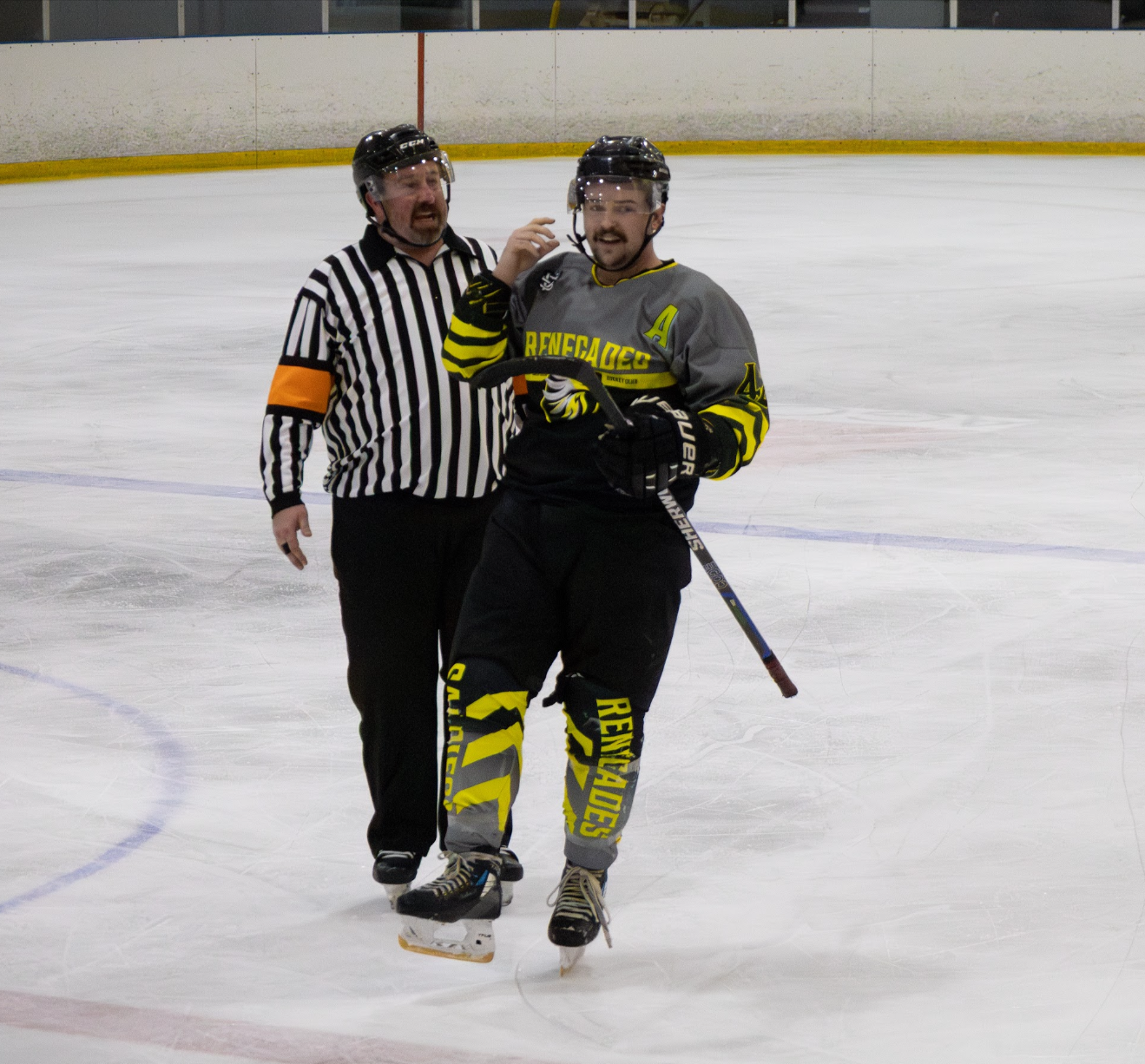An ice hockey player with a gray and black and yellow uniform and a referee in a black-and-white striped shirt on an ice rink.