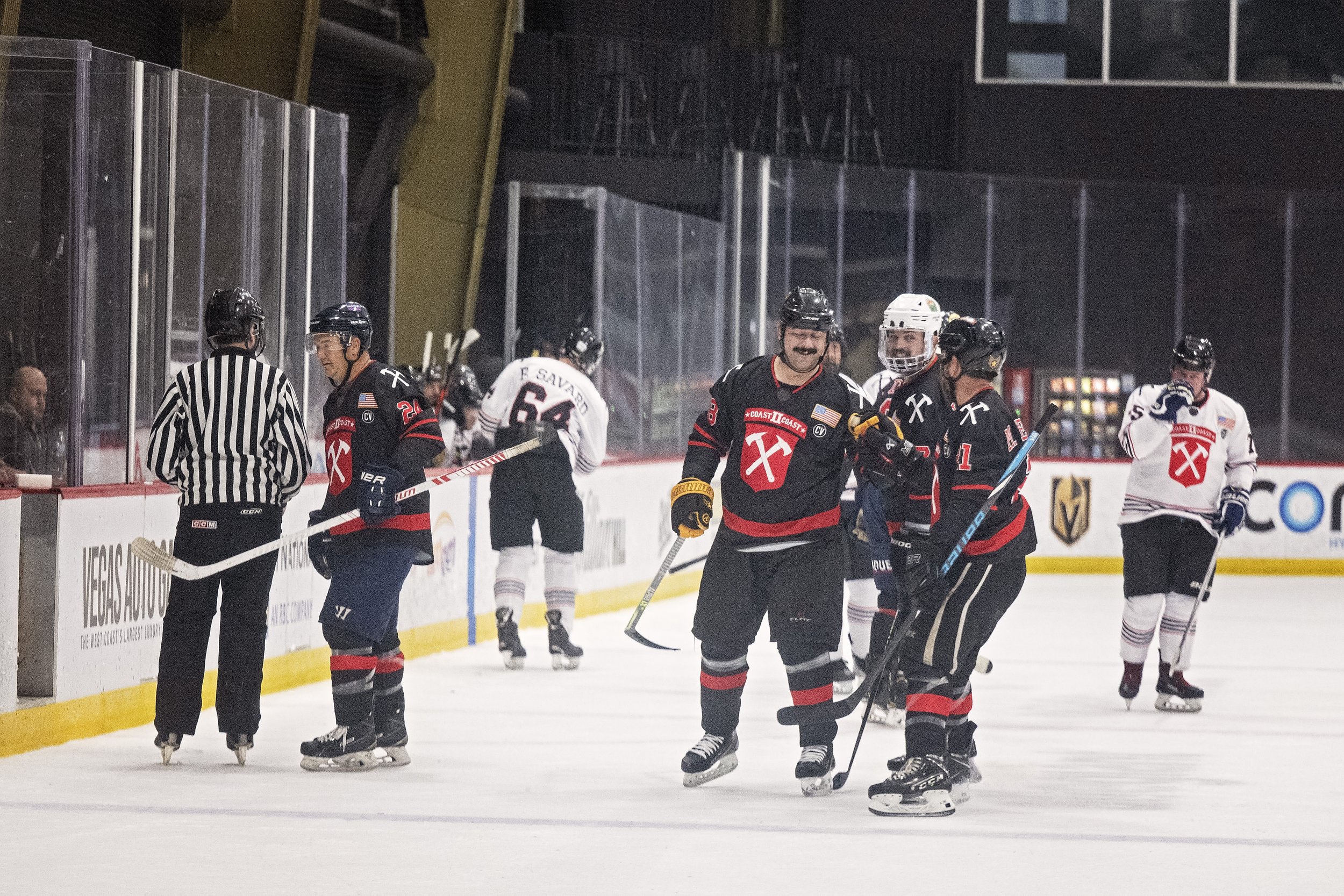 Hockey players on ice rink, some in black jerseys celebrating, others in white jerseys, with referee nearby.