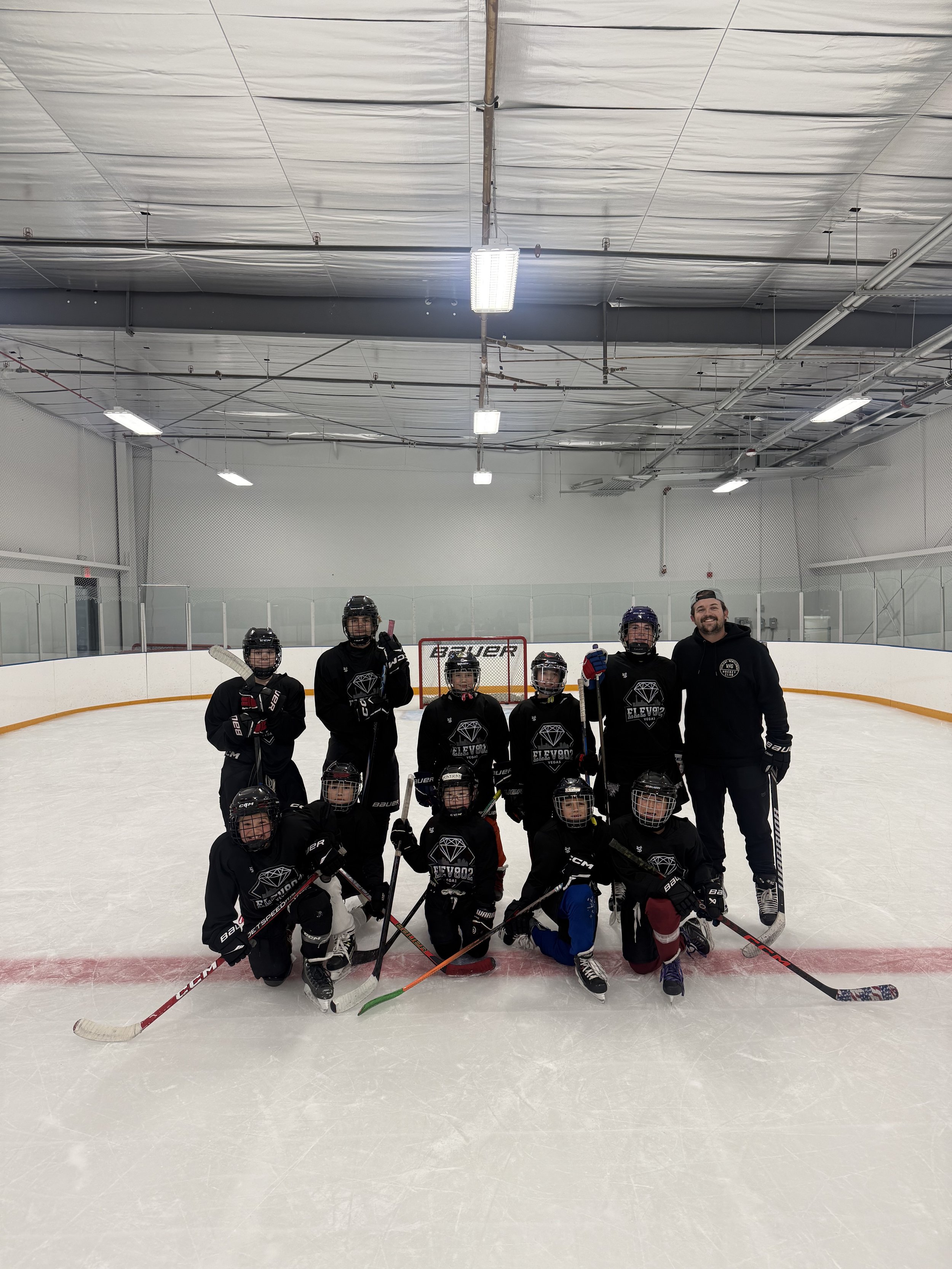 Kids and coach on an ice hockey rink with a hockey goal in the background, the kids are wearing hockey gear and holding hockey sticks.