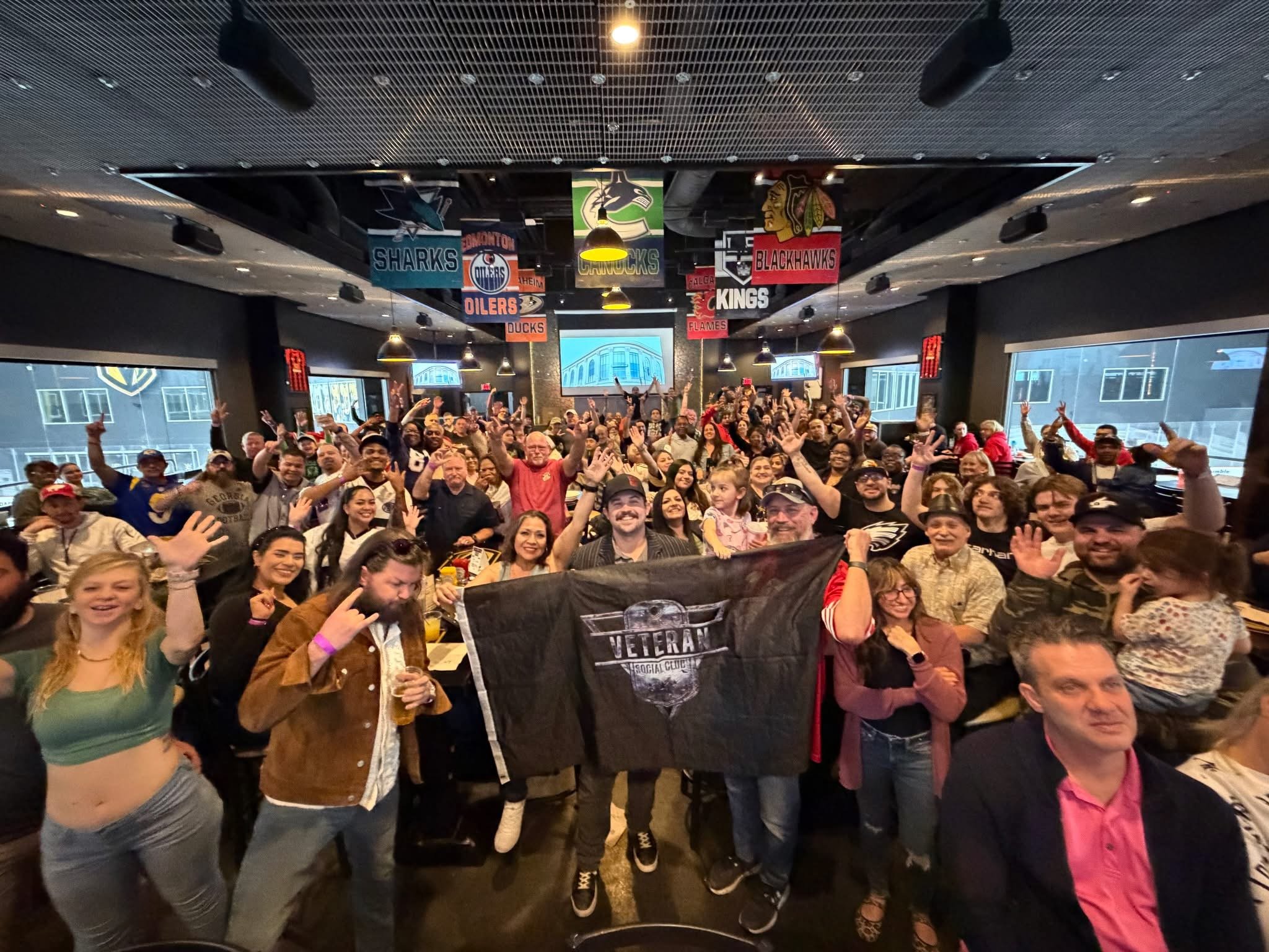 A large group of diverse people gathered in a social club or bar, many smiling and raising their hands. Some are holding a flag that says 'Veteran Social Club.' The background features banners of various hockey teams and large windows showing buildin