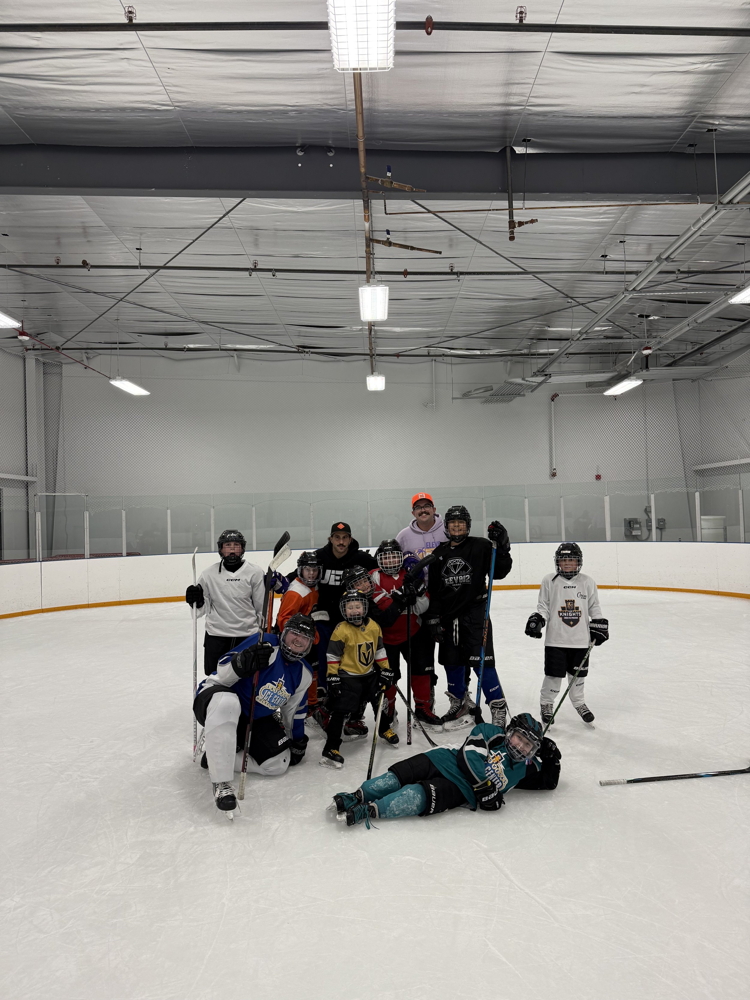 A group of young hockey players and coaches on an ice rink, posing for a photo after a game or practice, with some holding hockey sticks and wearing helmets, in an indoor ice rink.