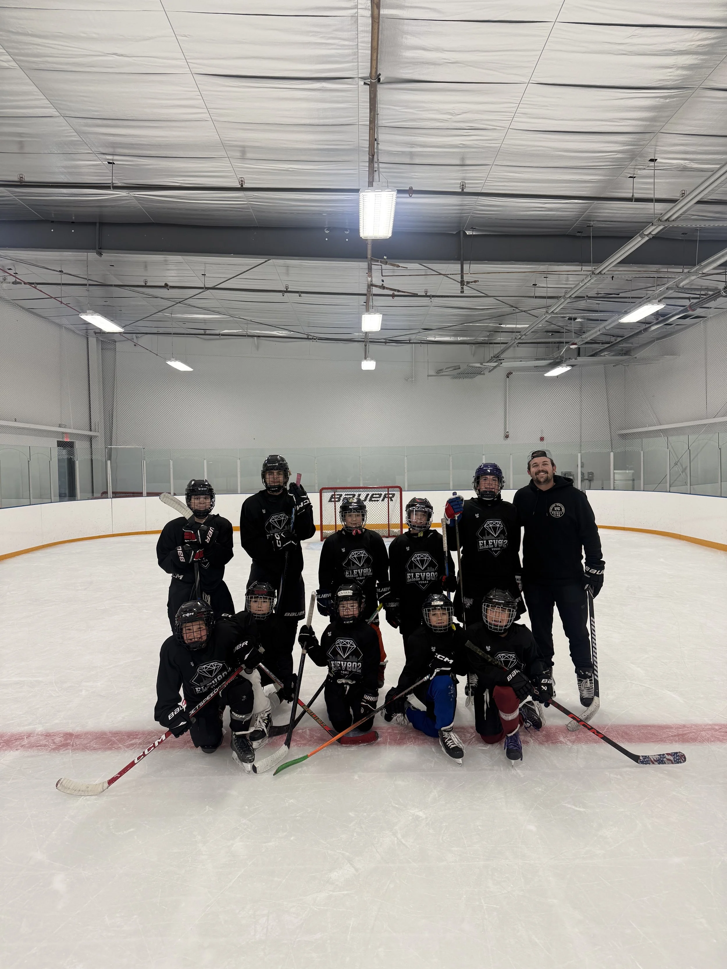 A youth hockey team with their coach posing on an ice rink, all wearing black jerseys and hockey gear, with a goalie in the back near the net.