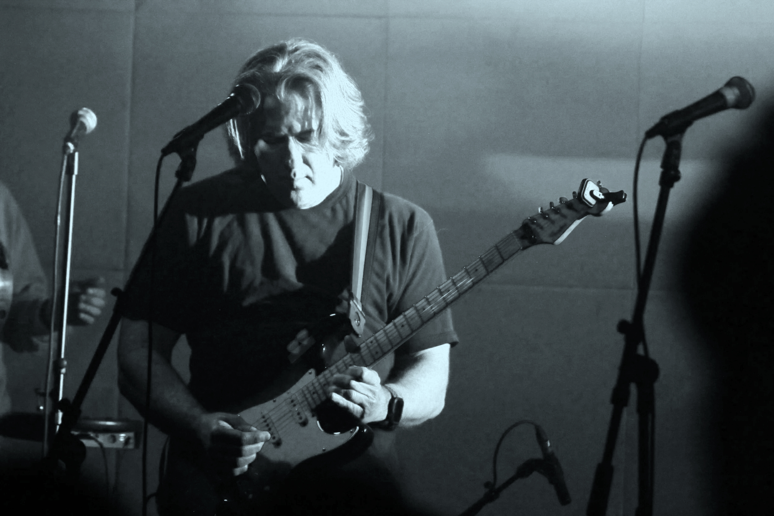 A man with shoulder-length hair playing an electric guitar on stage, surrounded by microphone stands, in a dimly lit setting.