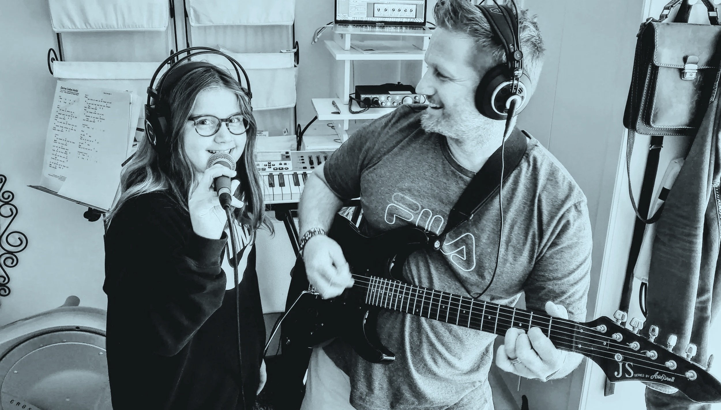 A young girl with glasses holding a microphone and singing, and a man playing an electric guitar, both wearing headphones, in a music room with instruments and music sheets.