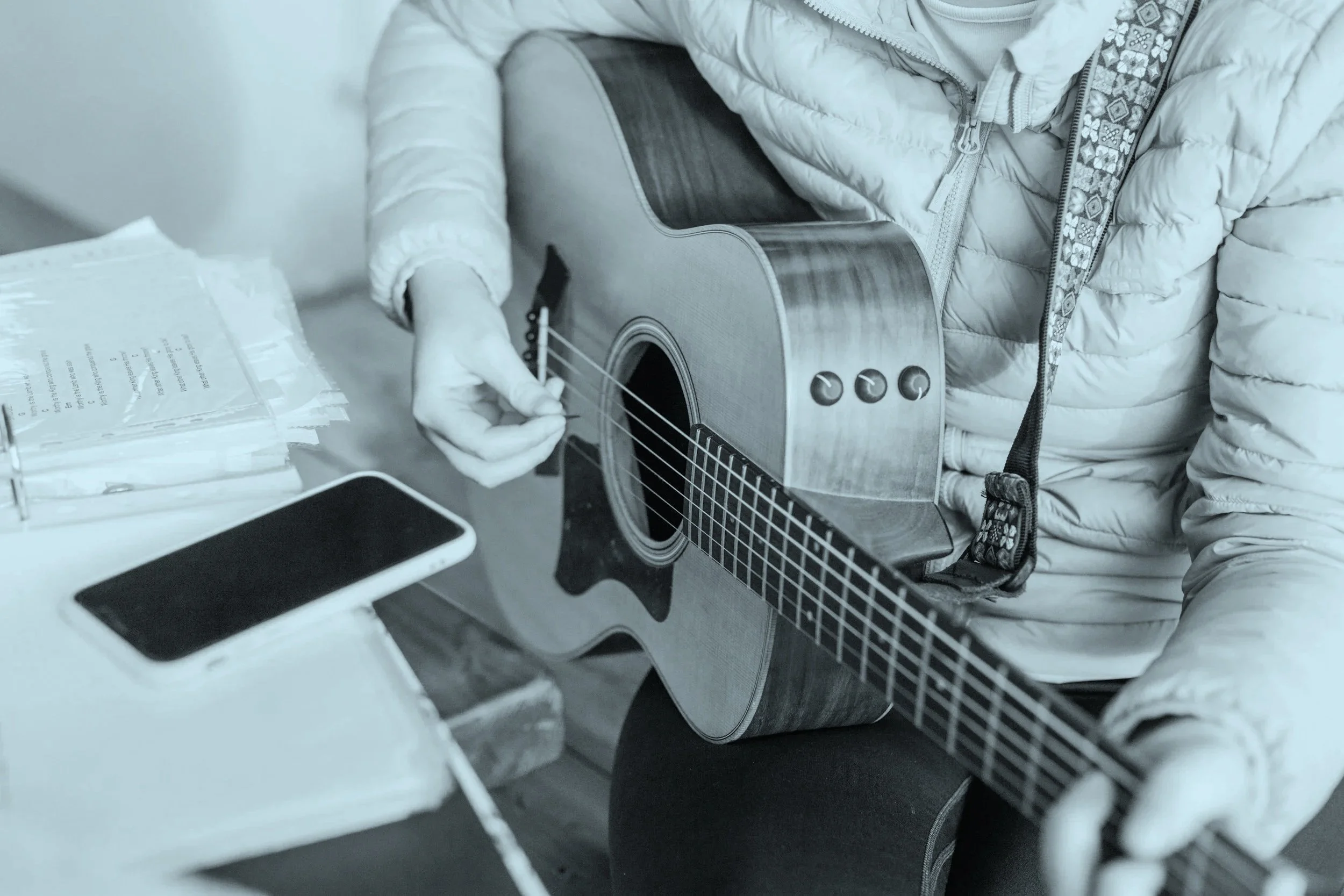 Person sitting at a table playing an acoustic guitar with a floral strap, a smartphone, and some papers.