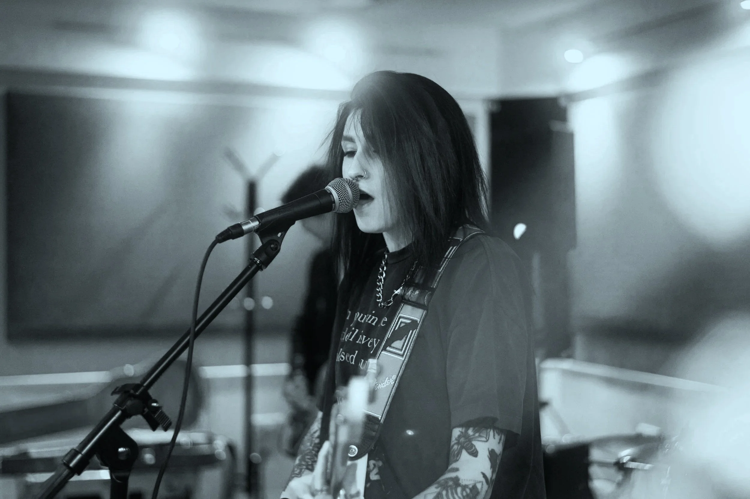 A woman with shoulder-length dark hair singing into a microphone in a music studio, playing an electric guitar, with tattoos visible on her arms.