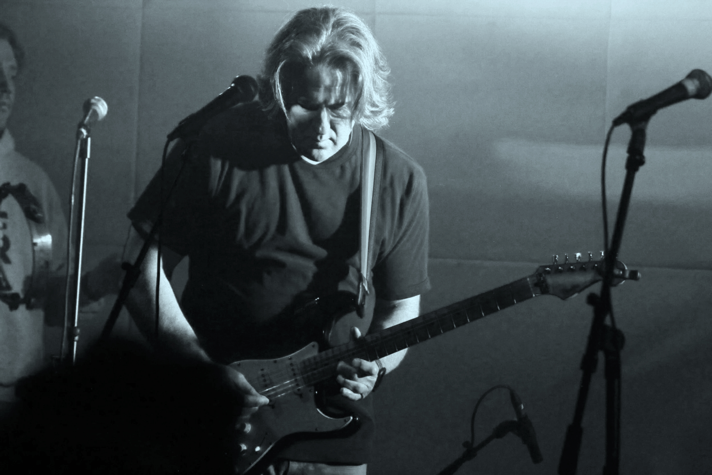 A person playing an electric guitar on stage, with a microphone stand nearby, in a black and white photograph.