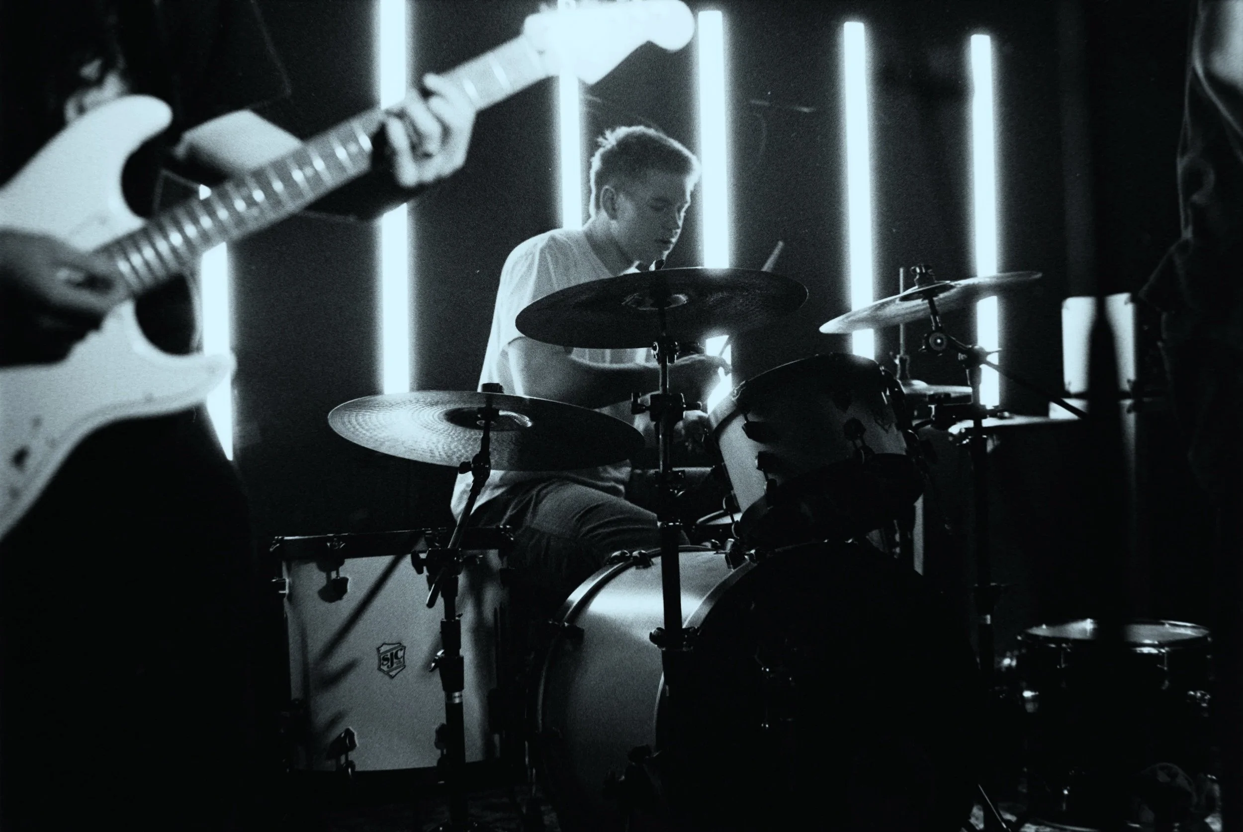 A black and white photograph of a band performing on stage, featuring a drummer and guitarists, with neon lights in the background.