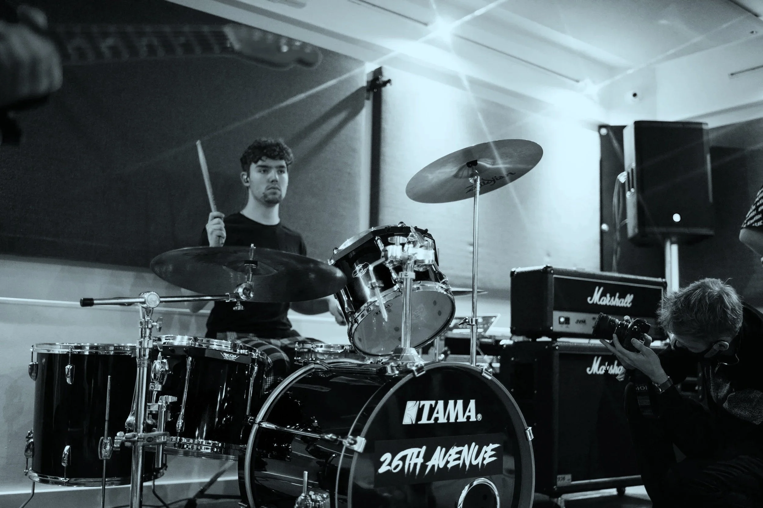 A young man sits behind a drum set, holding drumsticks in a music studio with amplifiers and speakers, as another person takes photos.