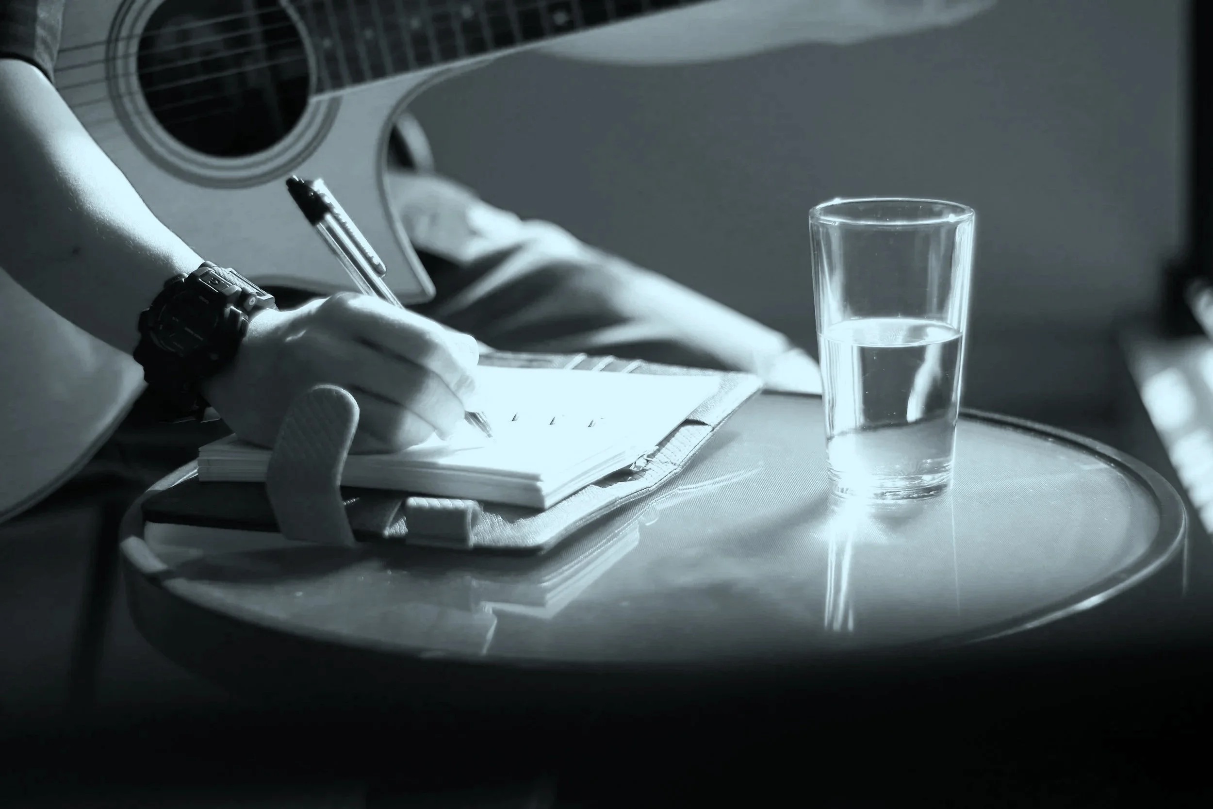 A person writing in a notebook with a pen, with a glass of water on a reflective table, a guitar partially visible in the background, in black and white.