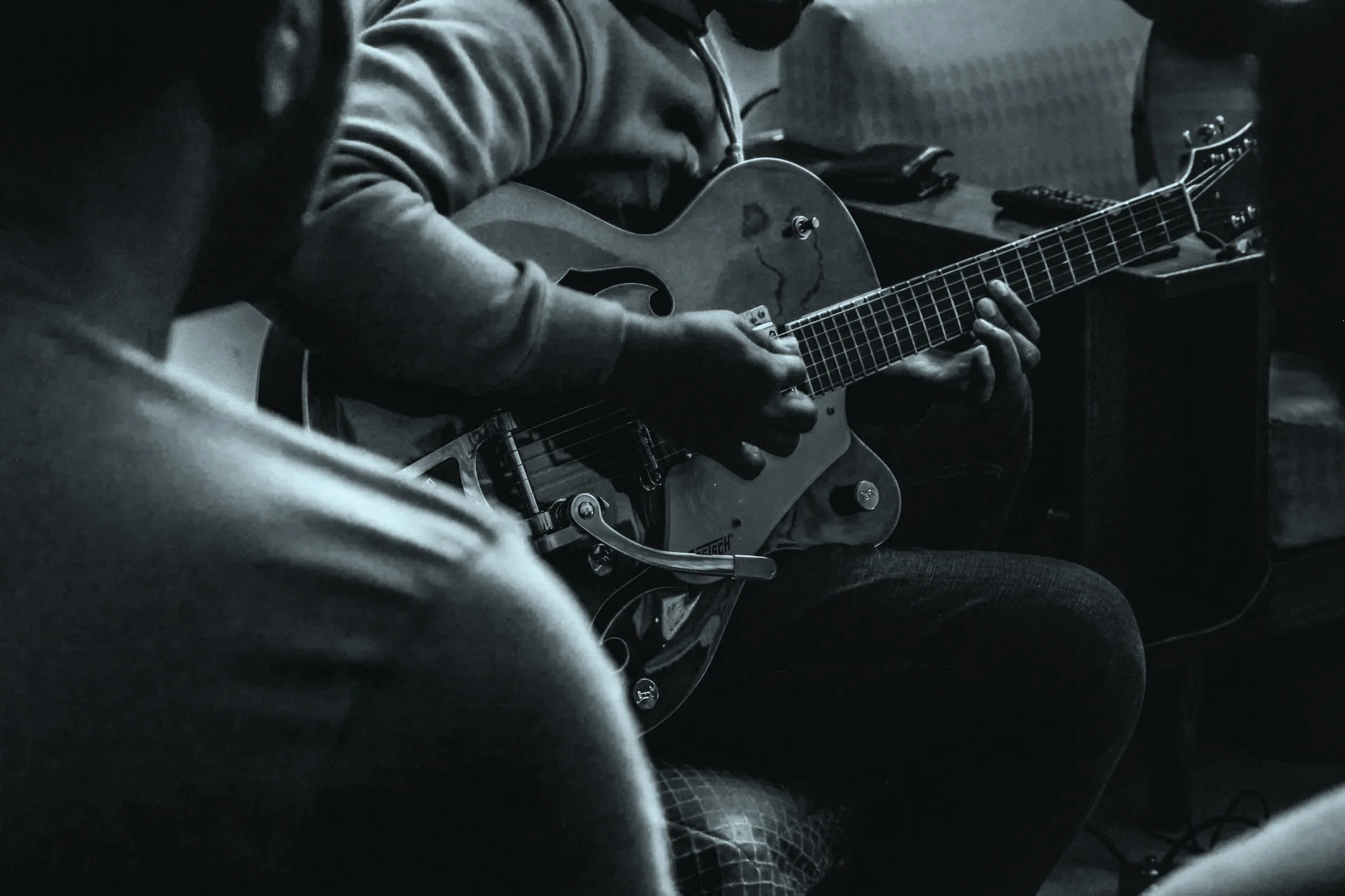 Person playing an electric guitar in a dimly lit room, with focus on their hands and the guitar.
