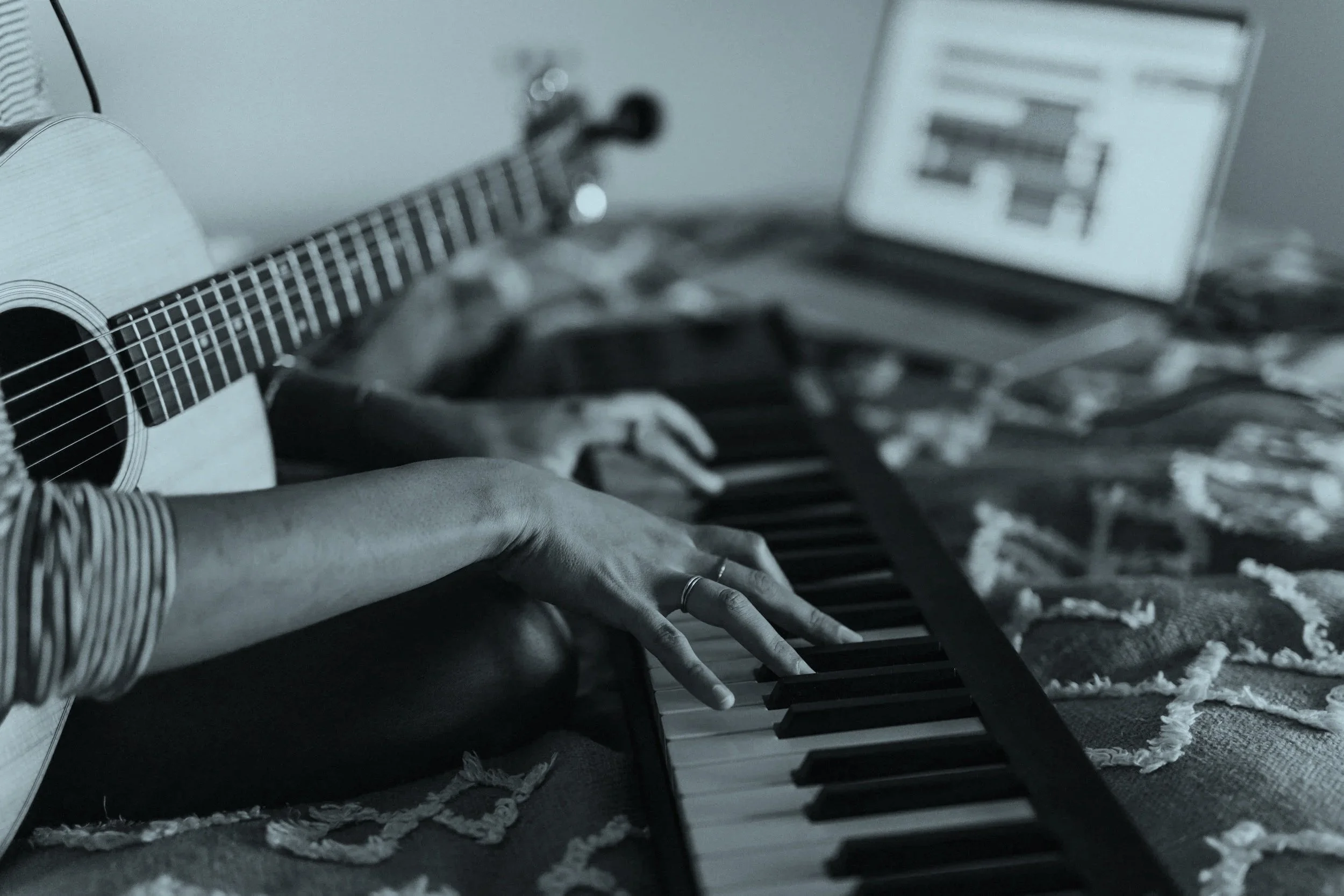 Person playing a keyboard and an acoustic guitar, with music software on a laptop in the background.