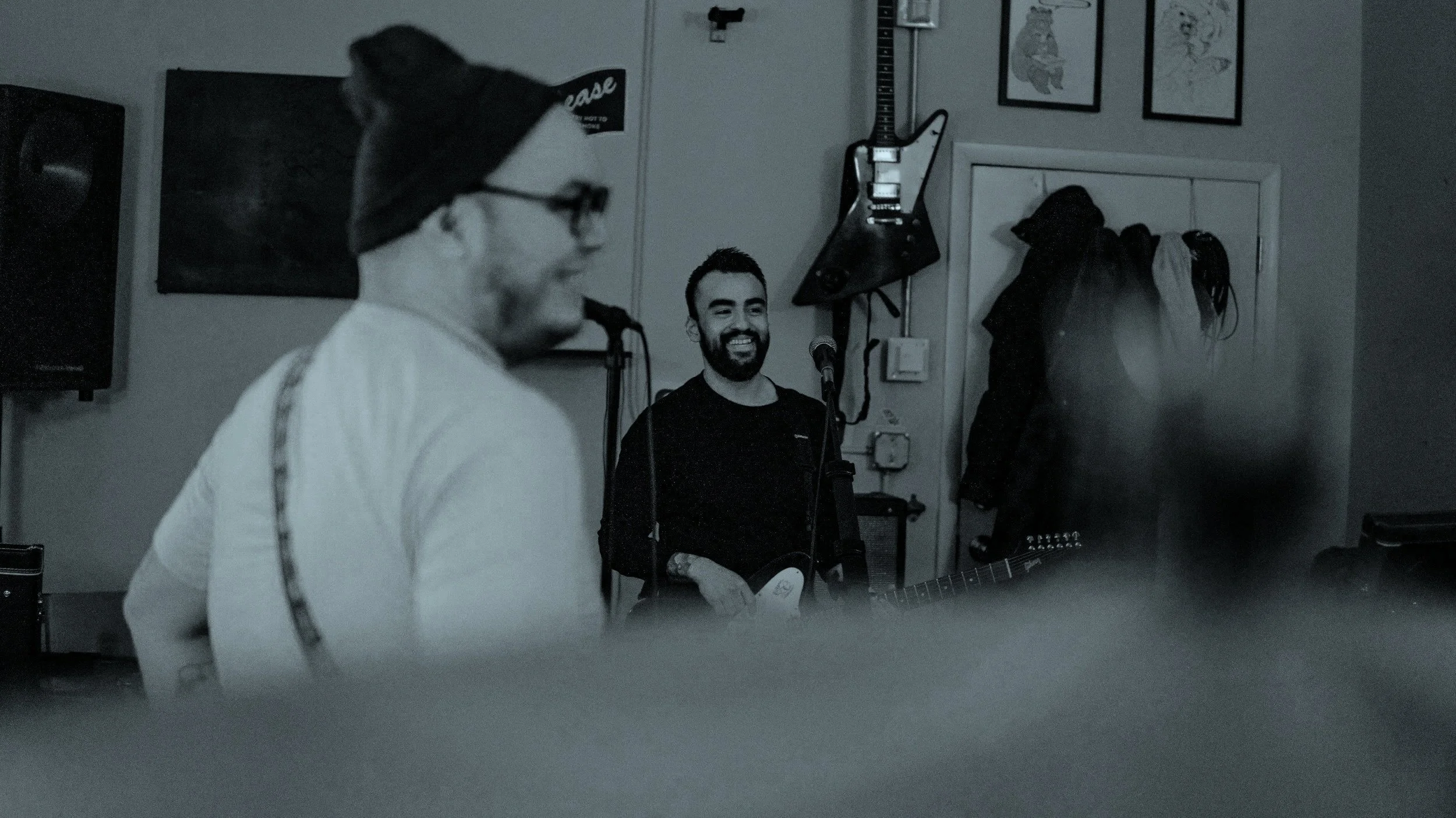 Two men in a music studio, one with a guitar smiling, and the other wearing glasses and a beanie, blurred in the foreground.