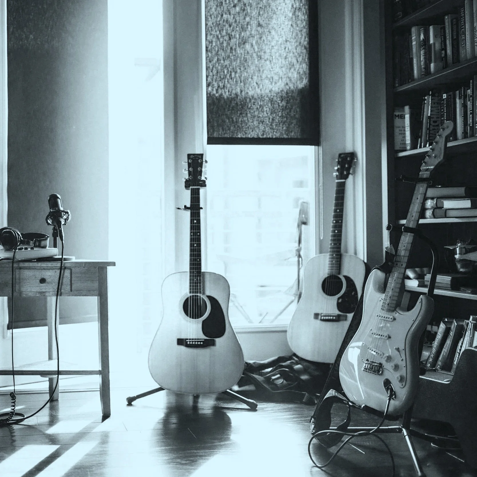 Three acoustic and electric guitars in a music room next to a window with bookshelves, a desk with headphones and a microphone.