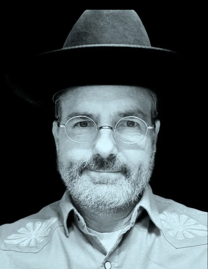 A black and white photo of a man wearing glasses, a hat, and a shirt with floral embroidery on the shoulders, smiling at the camera.