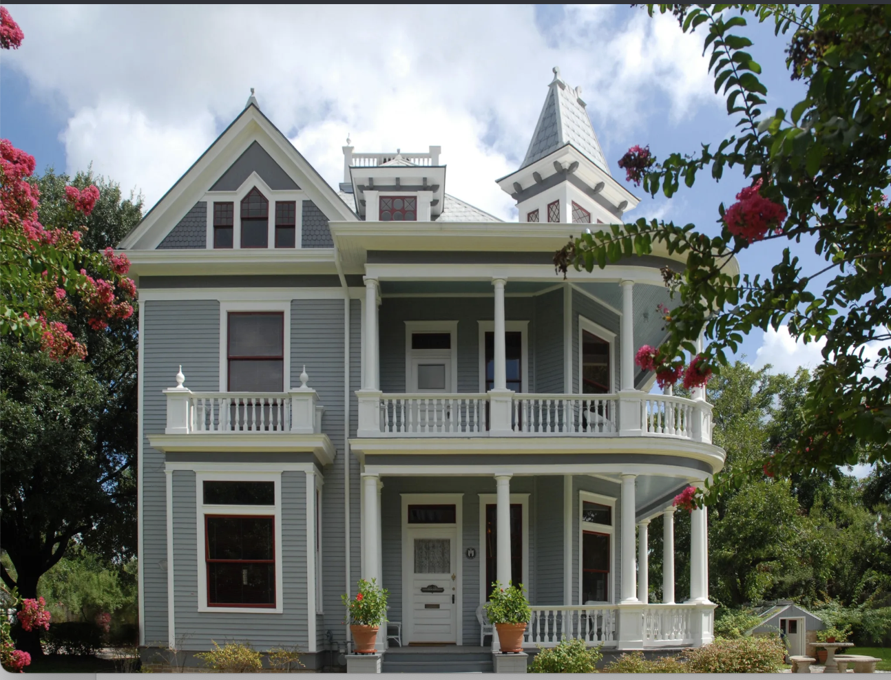 A large Victorian-style house with a gray exterior, white trim, and multiple porches, surrounded by trees and flowering plants.