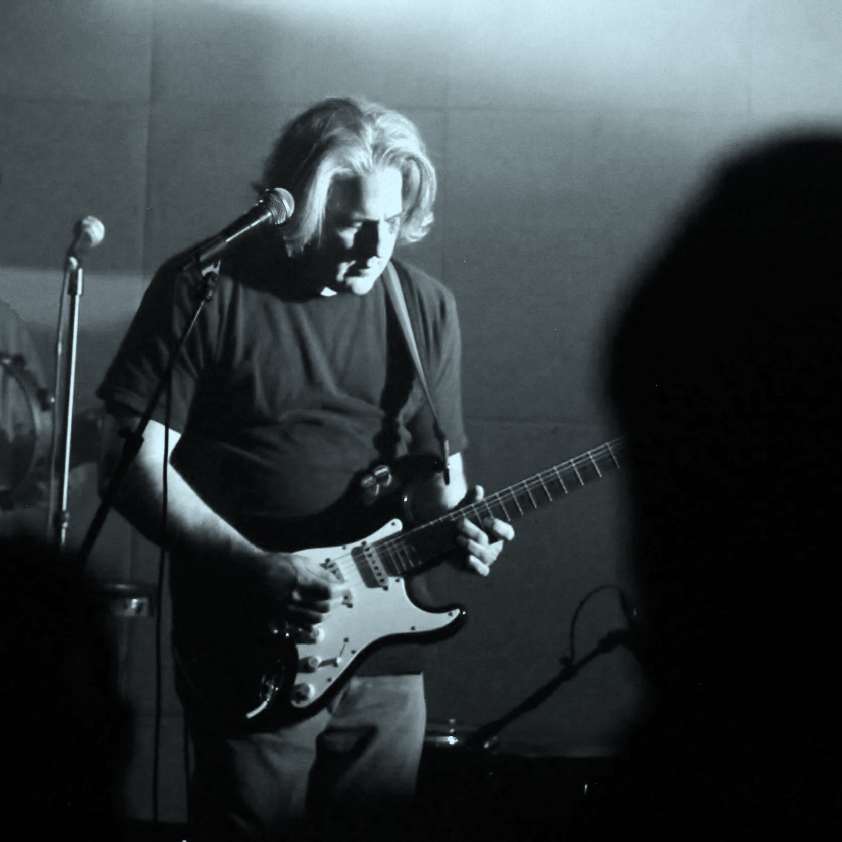 A person playing an electric guitar during a live performance in a dark venue, with a microphone stand nearby and another band member partially visible in the background.